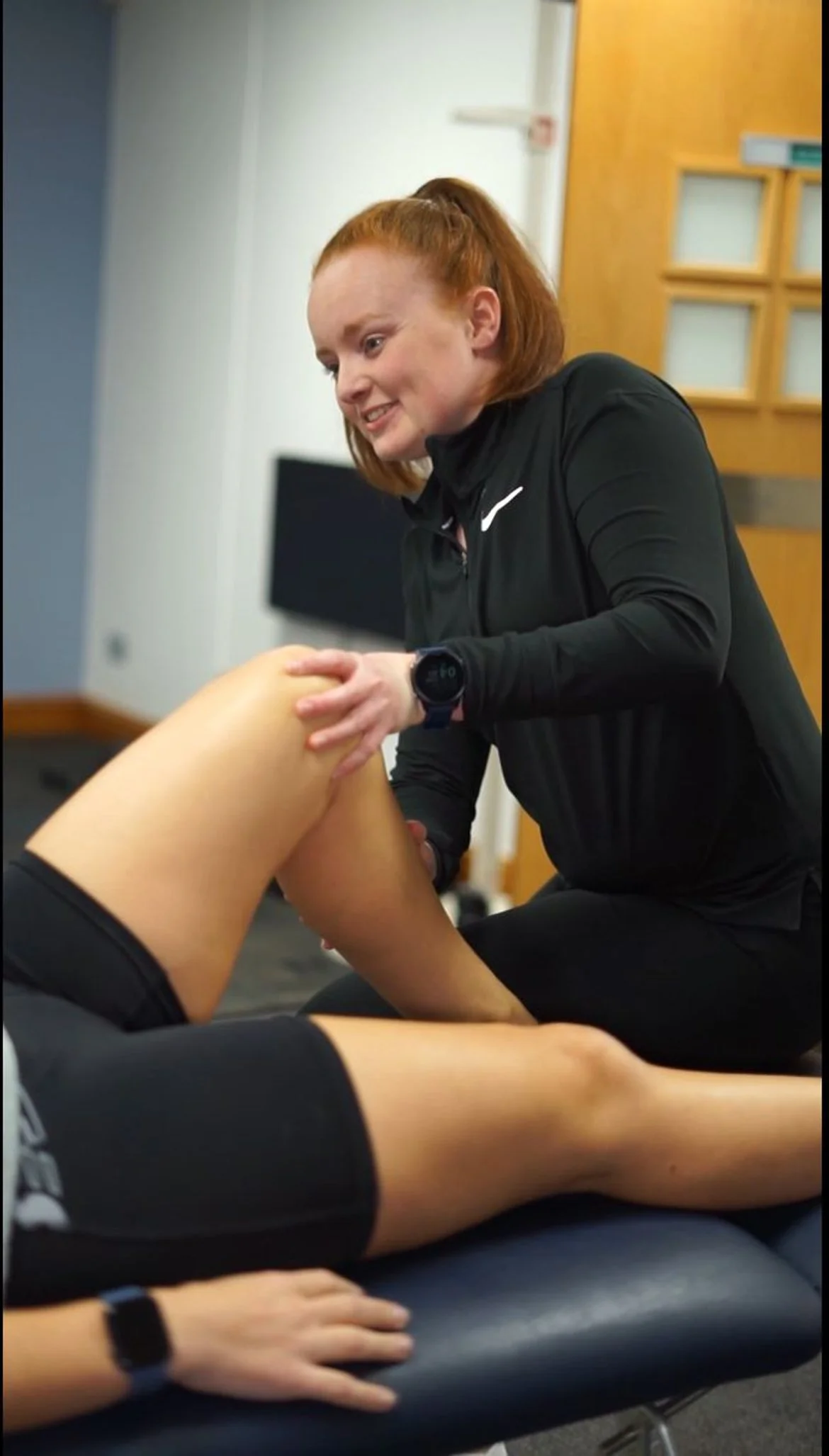 A woman wearing a black sports jacket is providing physical therapy or massage to a person's leg, which is lying on a black treatment table.
