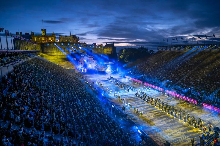 Full cast finale at the Royal Edinburgh Military Tattoo with massed performers, dramatic lighting effects, and castle projections, viewed from the side-stand seating under night skies.