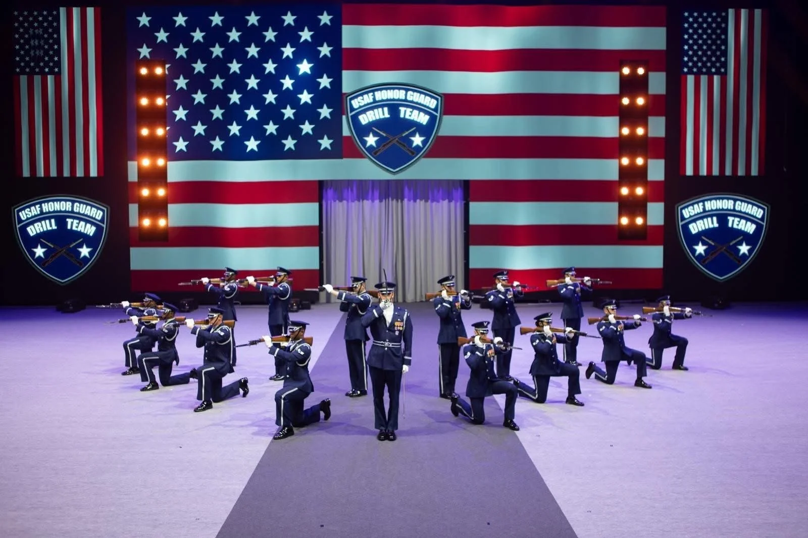 United States Air Force Honor Guard Drill Team performing a precision routine with rifles in front of a large American flag display at the Royal Edinburgh Military Tattoo.