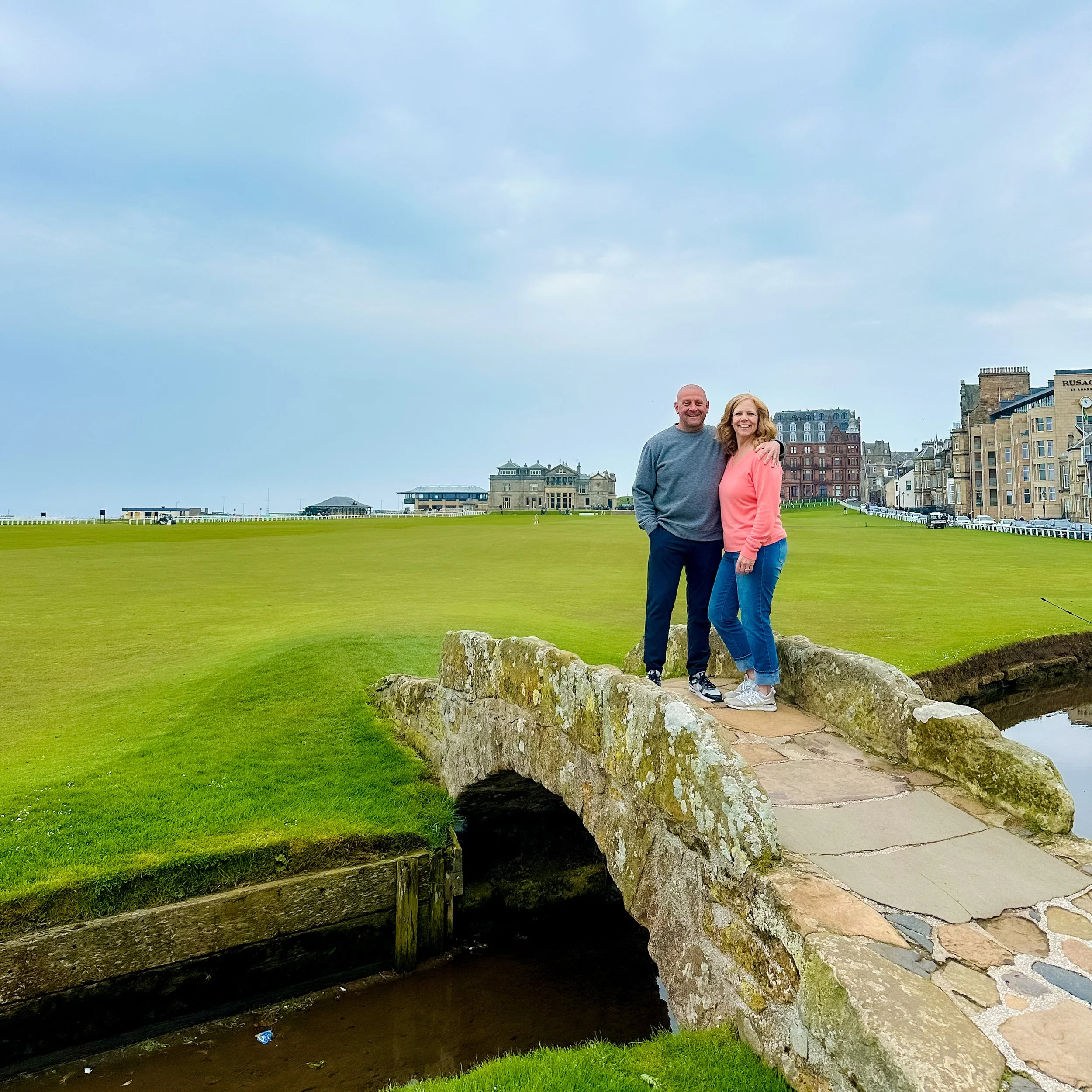 Two visitors standing on the historic Swilcan Bridge at the Old Course in St Andrews, Scotland, with the famous golf links and clubhouse buildings in the background.