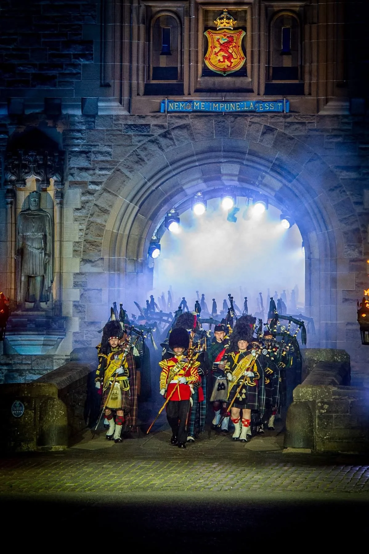 Massed pipe band in full ceremonial dress emerging through the illuminated gate of Edinburgh Castle at the Royal Edinburgh Military Tattoo, with mist and lights creating a dramatic atmosphere.