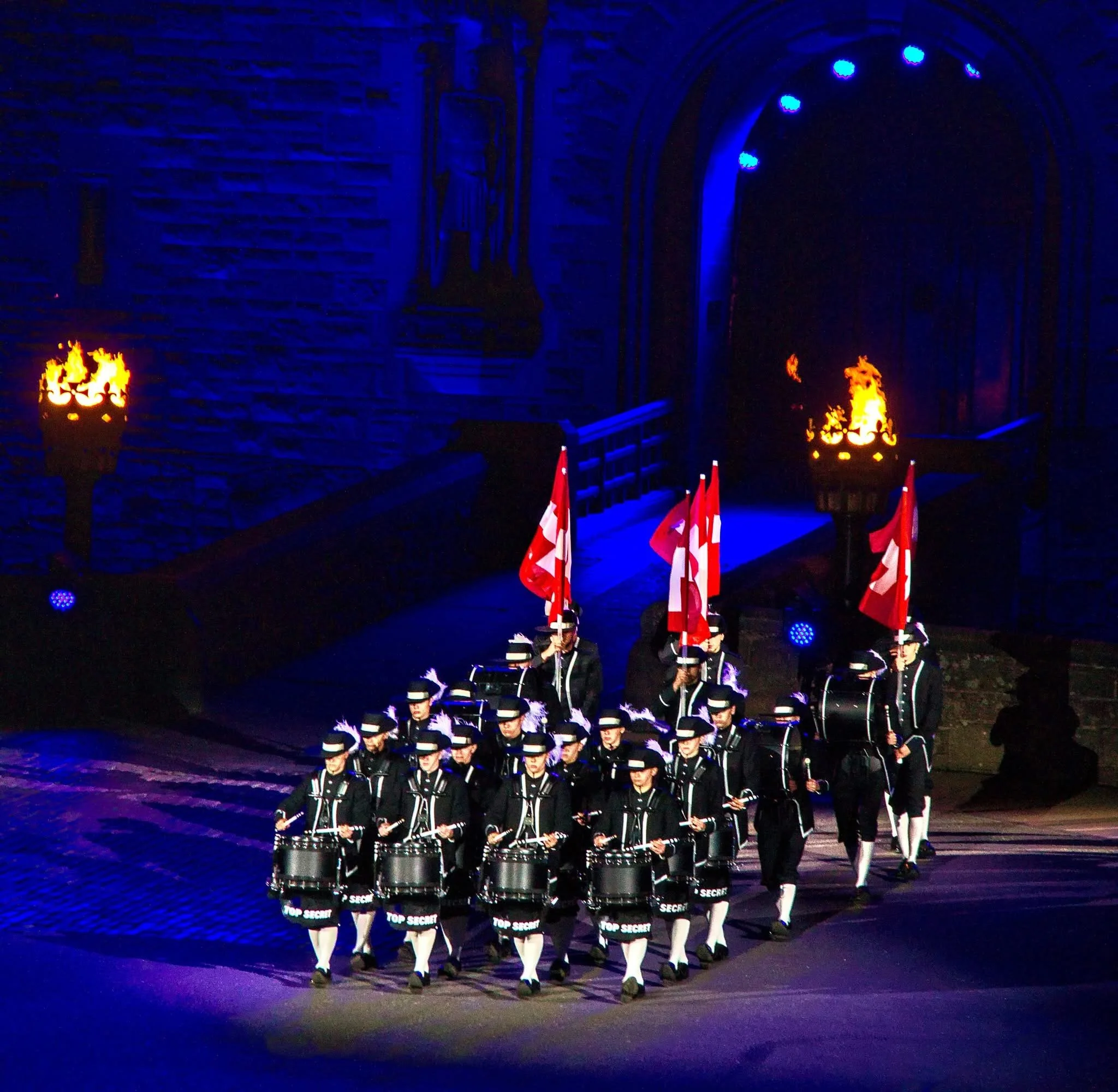 Top Secret Drum Corps from Switzerland performing at the Royal Edinburgh Military Tattoo, marching with drums and Swiss flags under torchlight at Edinburgh Castle.