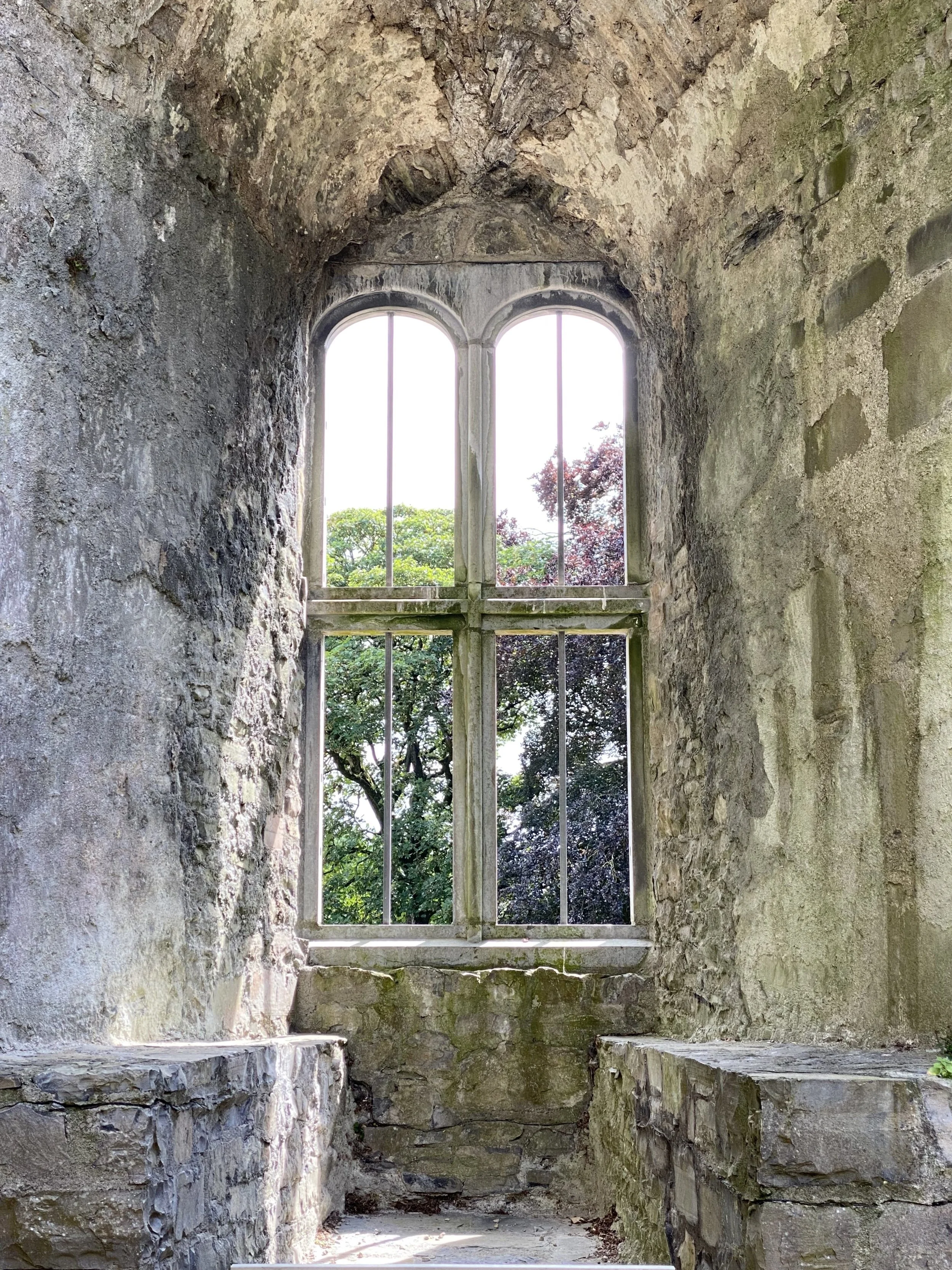 Old stone window with iron bars showing a view of trees outside.