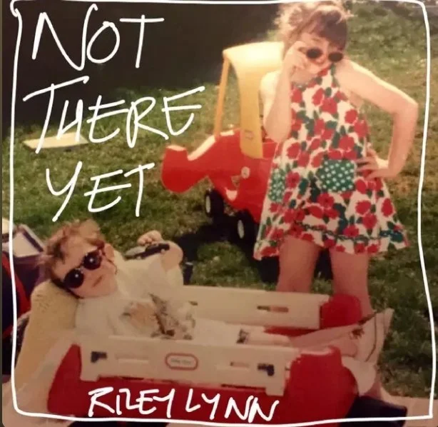 Two young girls enjoy a sunny day outdoors, one sitting in a toy wagon wearing sunglasses and a white shirt, the other standing next to her in a red, green, and white patterned dress, also wearing sunglasses. Text overlays say "Not There Yet" and "Riley Lynn."