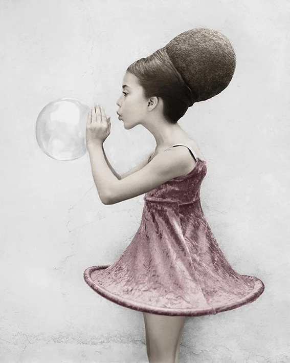 A young woman with an oversized textured bun hairstyle, wearing a pink velvet dress that flares out like a spinning top, is holding a soap bubble close to her face as if about to pop it, against a plain gray wall.