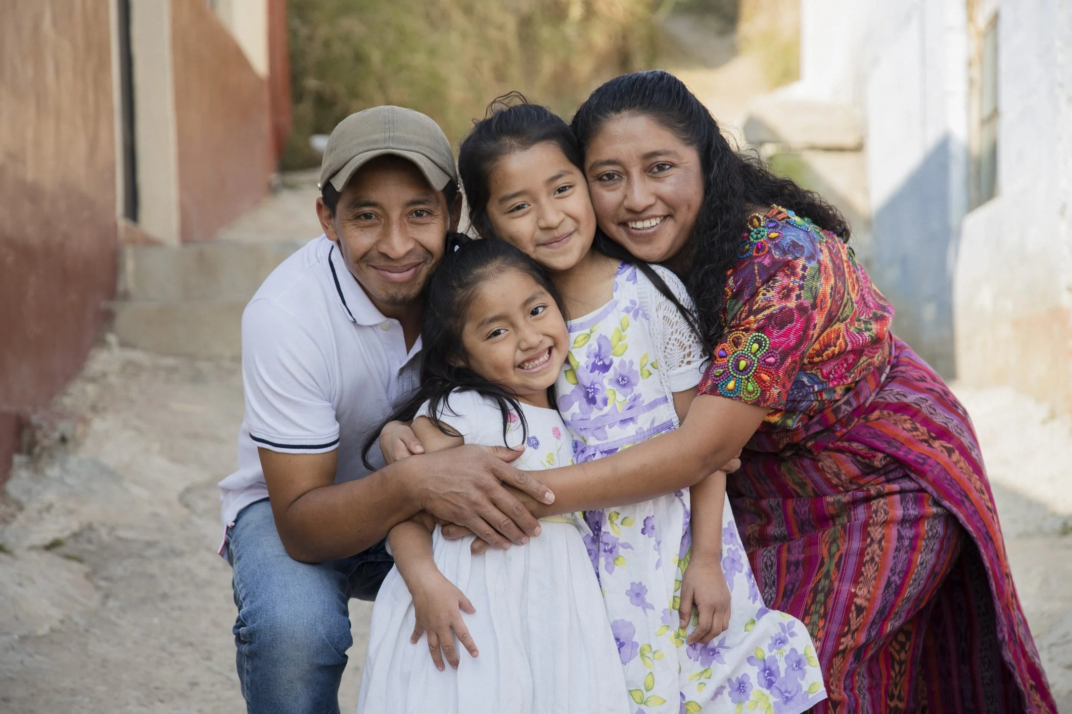 A smiling family of four, including a man, woman, and two young girls, outdoors on a pathway. The man wears a cap and white polo shirt, the woman is in a colorful patterned dress, and the girls wear floral dresses.