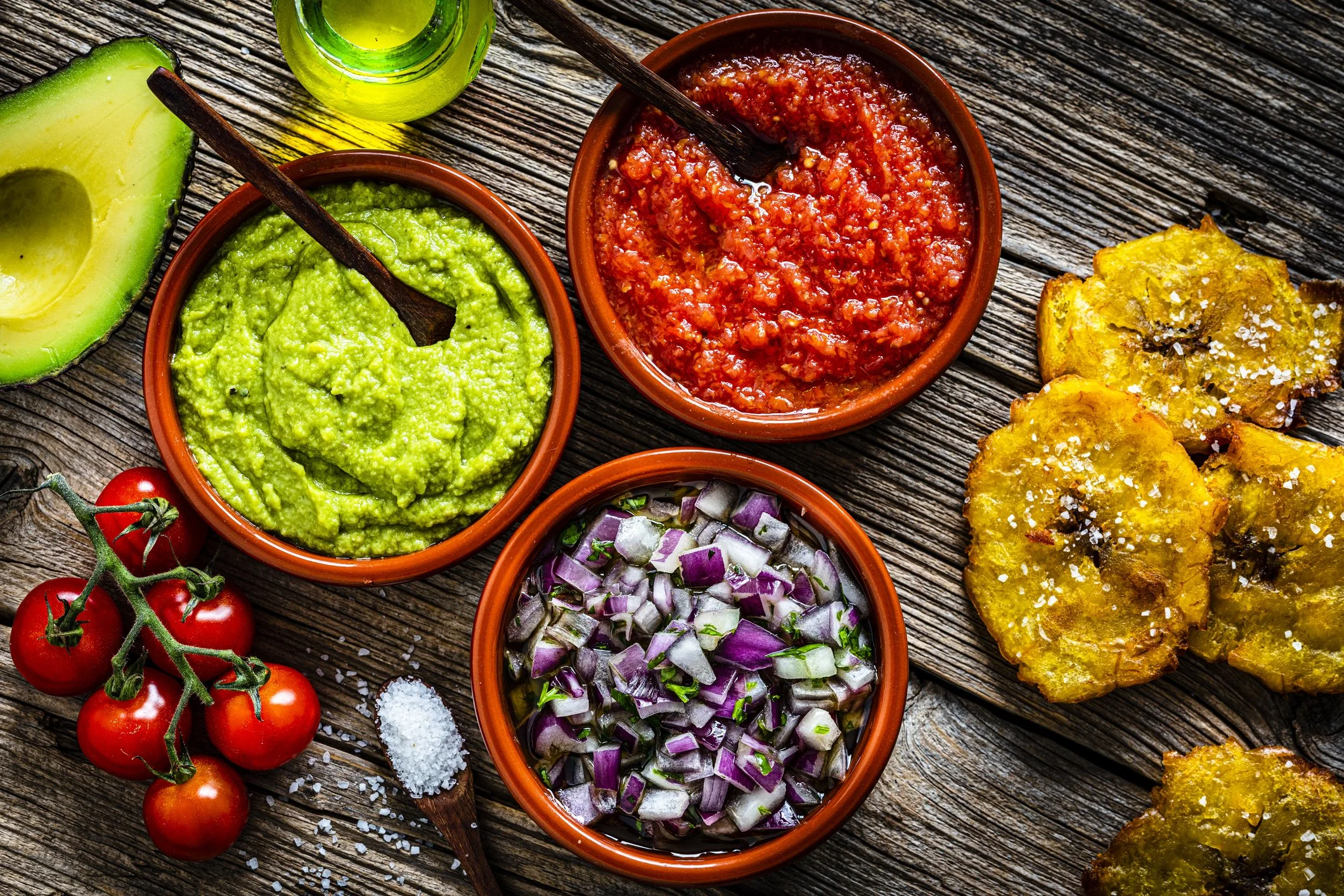 Assorted sauces and toppings including guacamole, tomato salsa, diced onions, cherry tomatoes, fried plantains, and rock salt on a rustic wooden table.