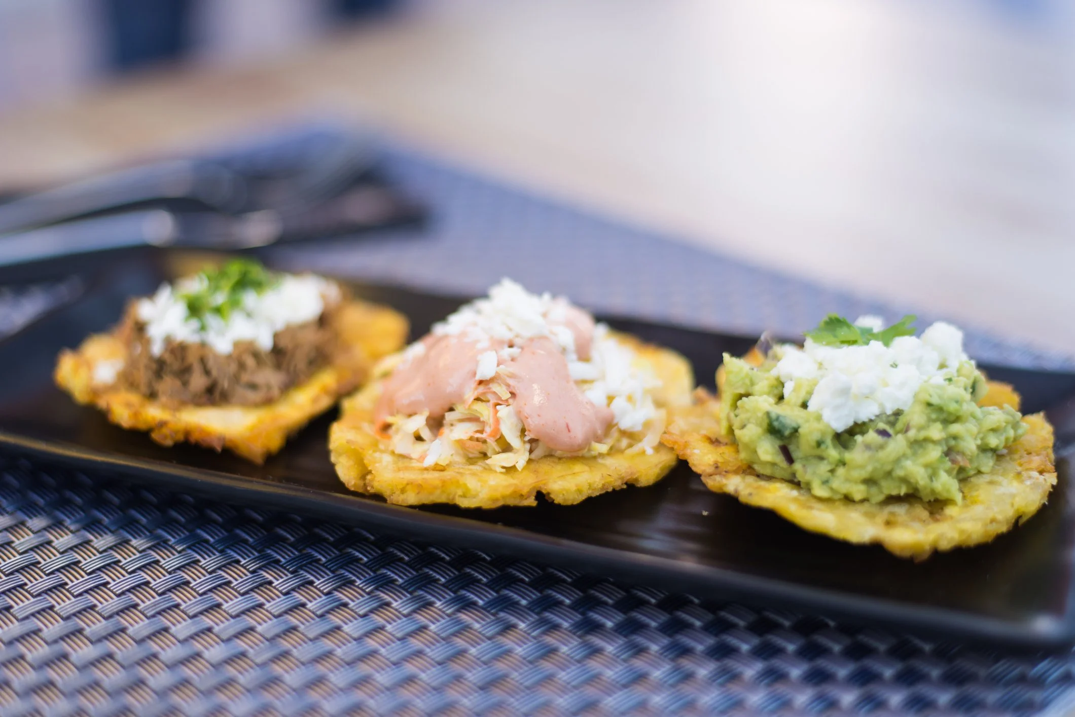 Three arepas on a black rectangular plate, topped with shredded meat, slaw with sauce, and guacamole with cheese on a textured placemat background.