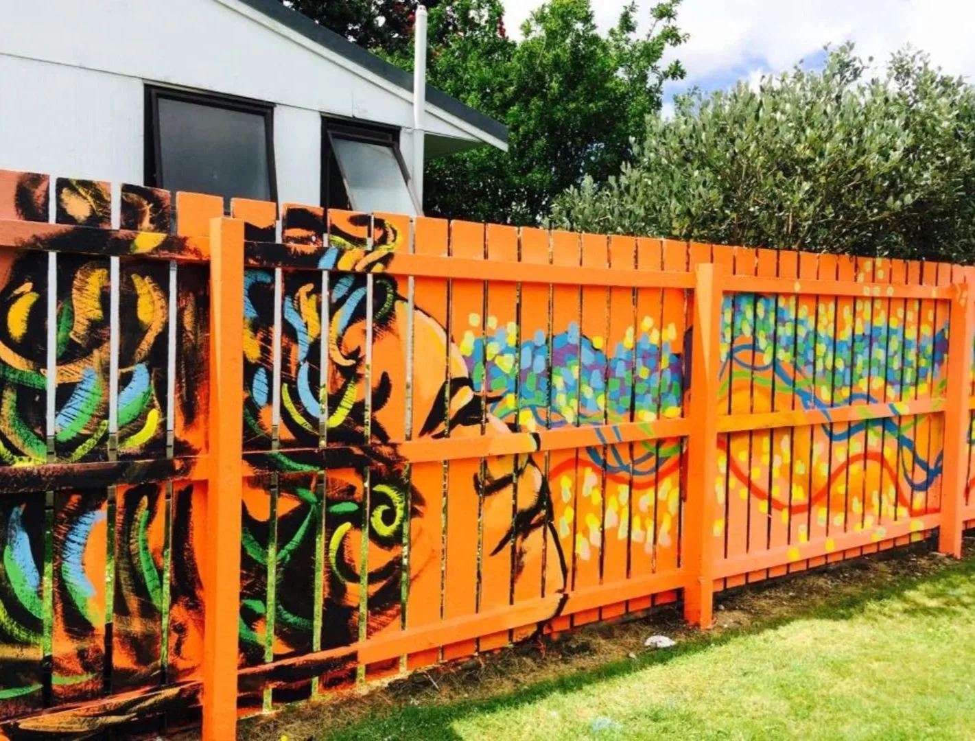 A colorful mural of abstract swirling patterns and flowers painted on a black-painted metal fence with orange frames, set in a grassy area with a house and green trees in the background.