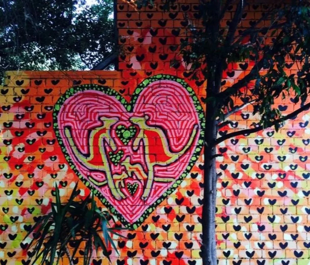 Colorful mural on a brick wall with a heart-shaped design, surrounded by smaller hearts, and a tree partially covering the artwork.