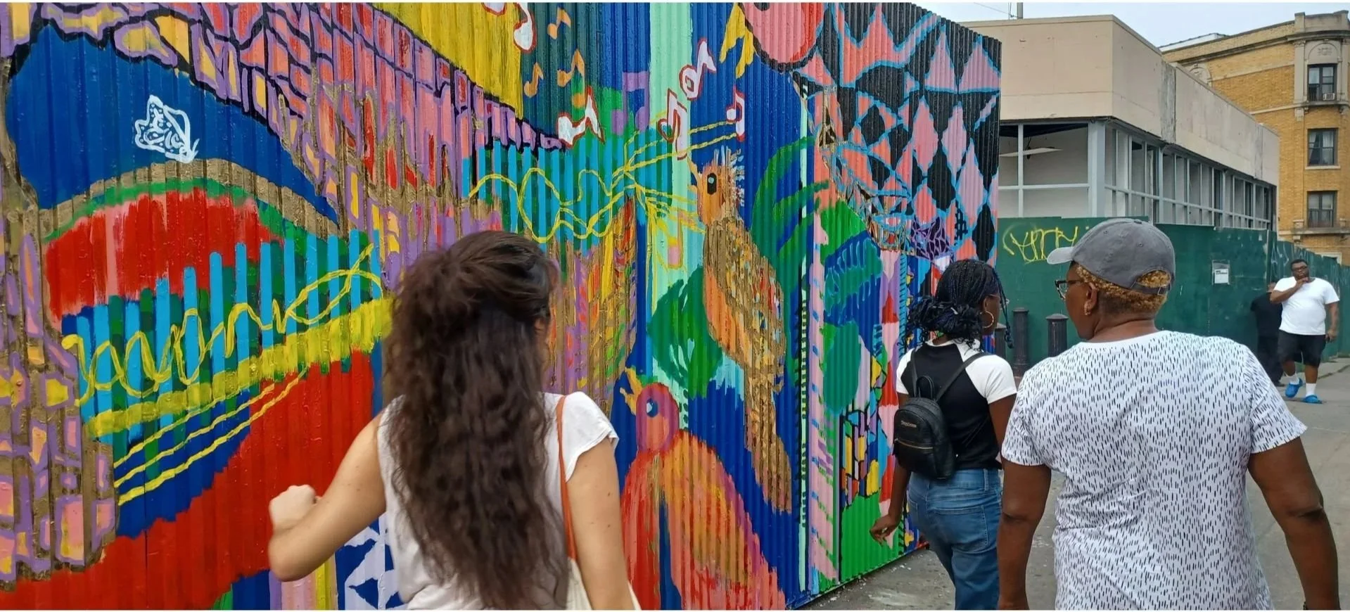 People viewing a colorful mural on a corrugated metal fence in an urban outdoor setting.