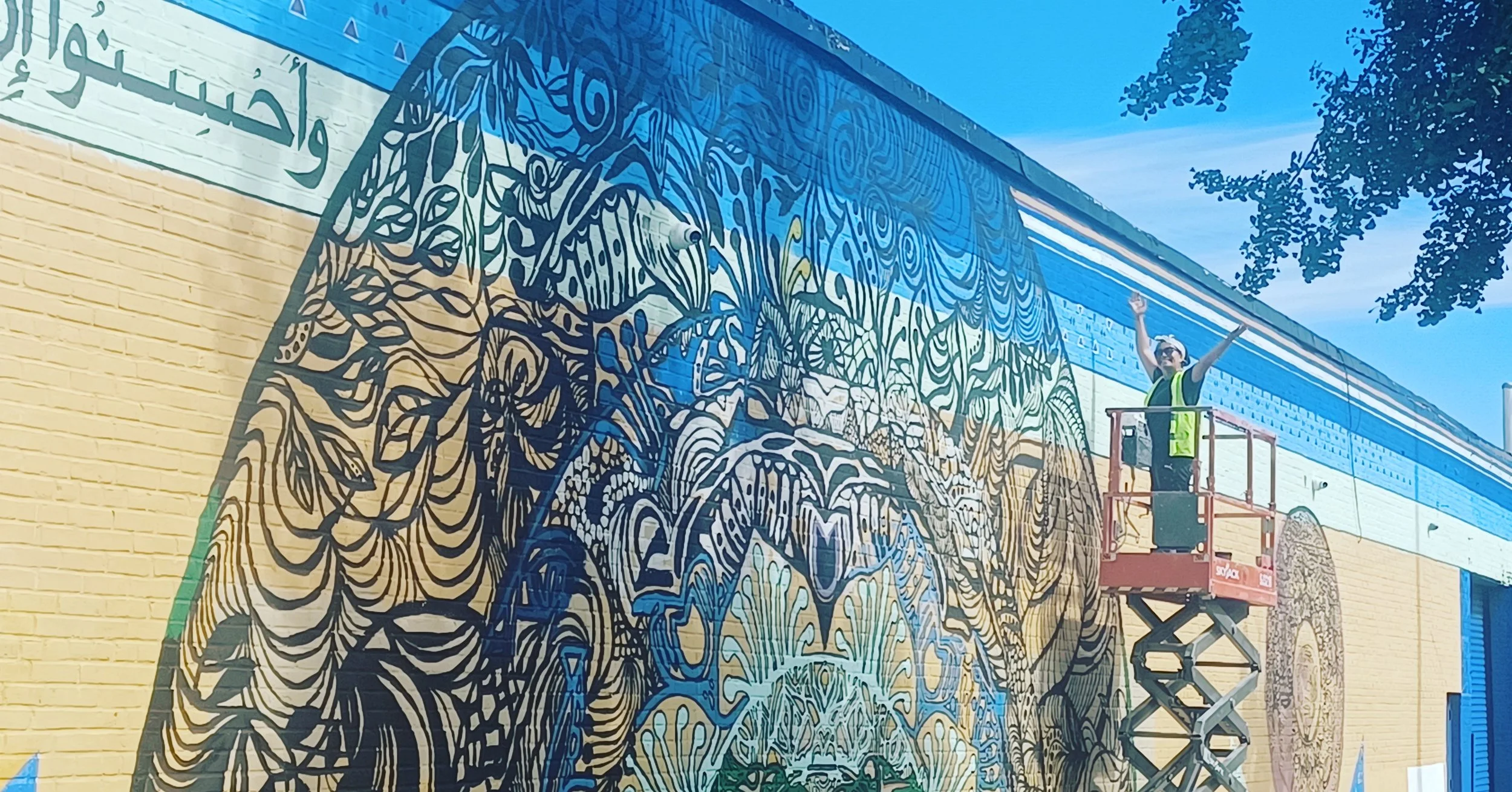 A man wearing a safety vest on a scissor lift painting a colorful mural on a brick wall, with trees and a blue sky overhead.