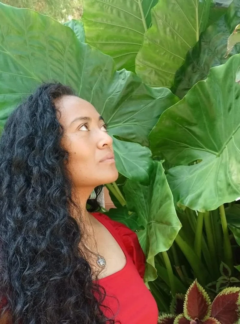 A woman with long, curly black hair looking up, standing next to large green leaves.