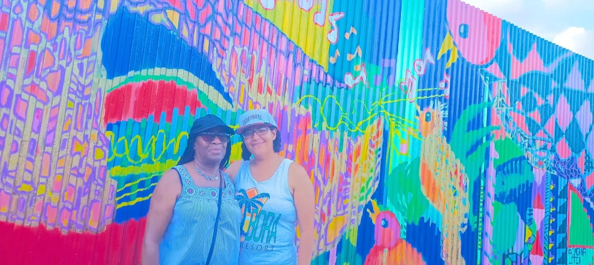 Two women standing in front of a colorful mural on a corrugated metal wall, wearing casual summer clothing, hats, and sunglasses, smiling at the camera.