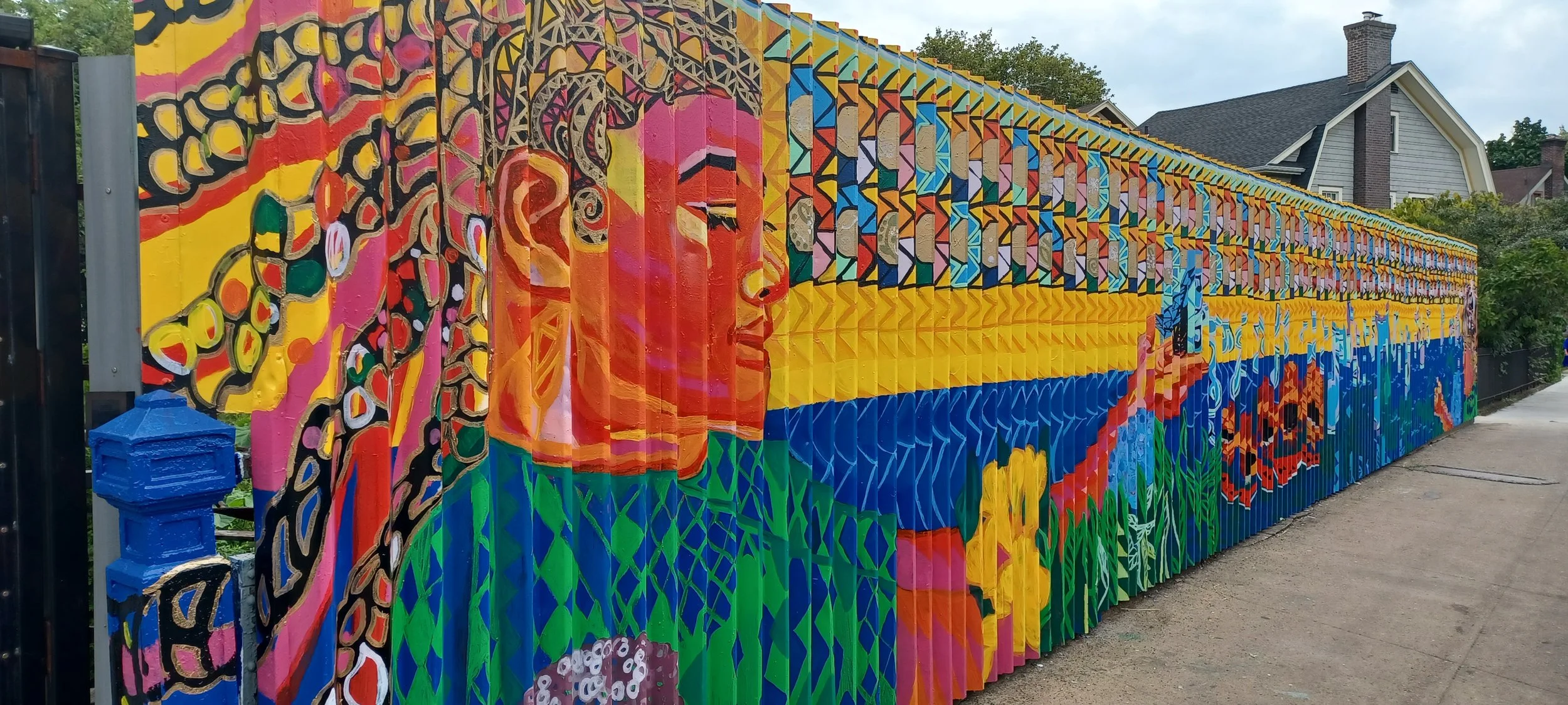 Colorful mural painting on a long fence depicting a woman with a blue headscarf, surrounded by vibrant colors and geometric patterns, with houses and trees in the background.