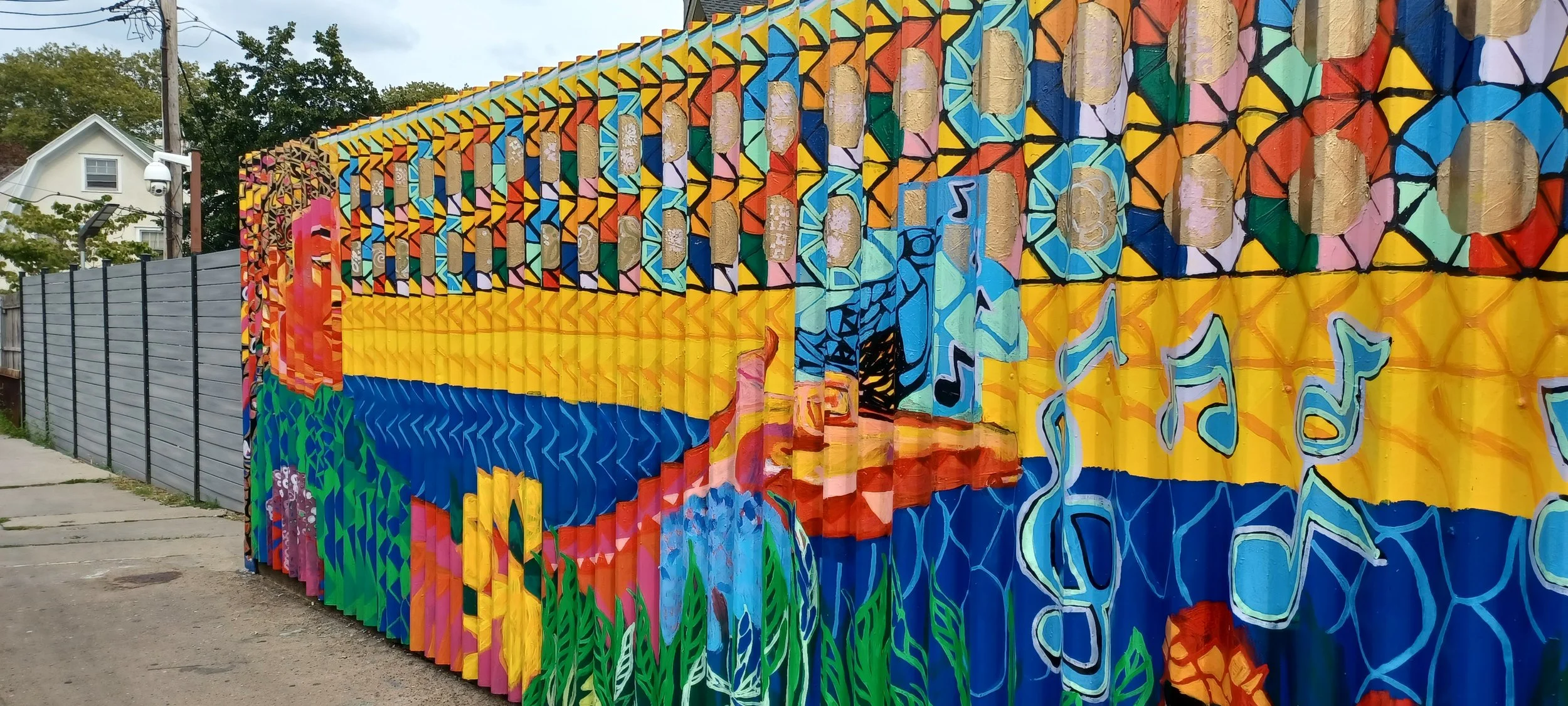 Colorful mural painted on a metal fence featuring abstract shapes, musical notes, and a sunset scene with water, mountains, and grass.
