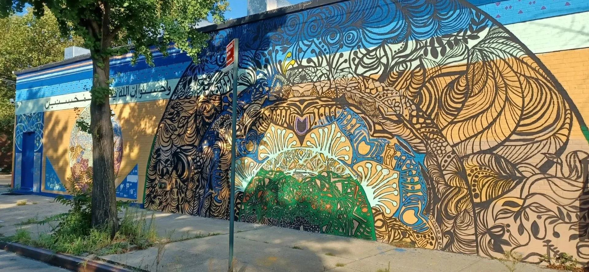 Colorful mural on a building wall with intricate abstract patterns and a large arch design, surrounded by a tree and sidewalk in an urban setting.