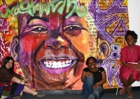 Three women sitting in front of a large colorful mural of a smiling person's face, with vibrant graffiti elements in the background.