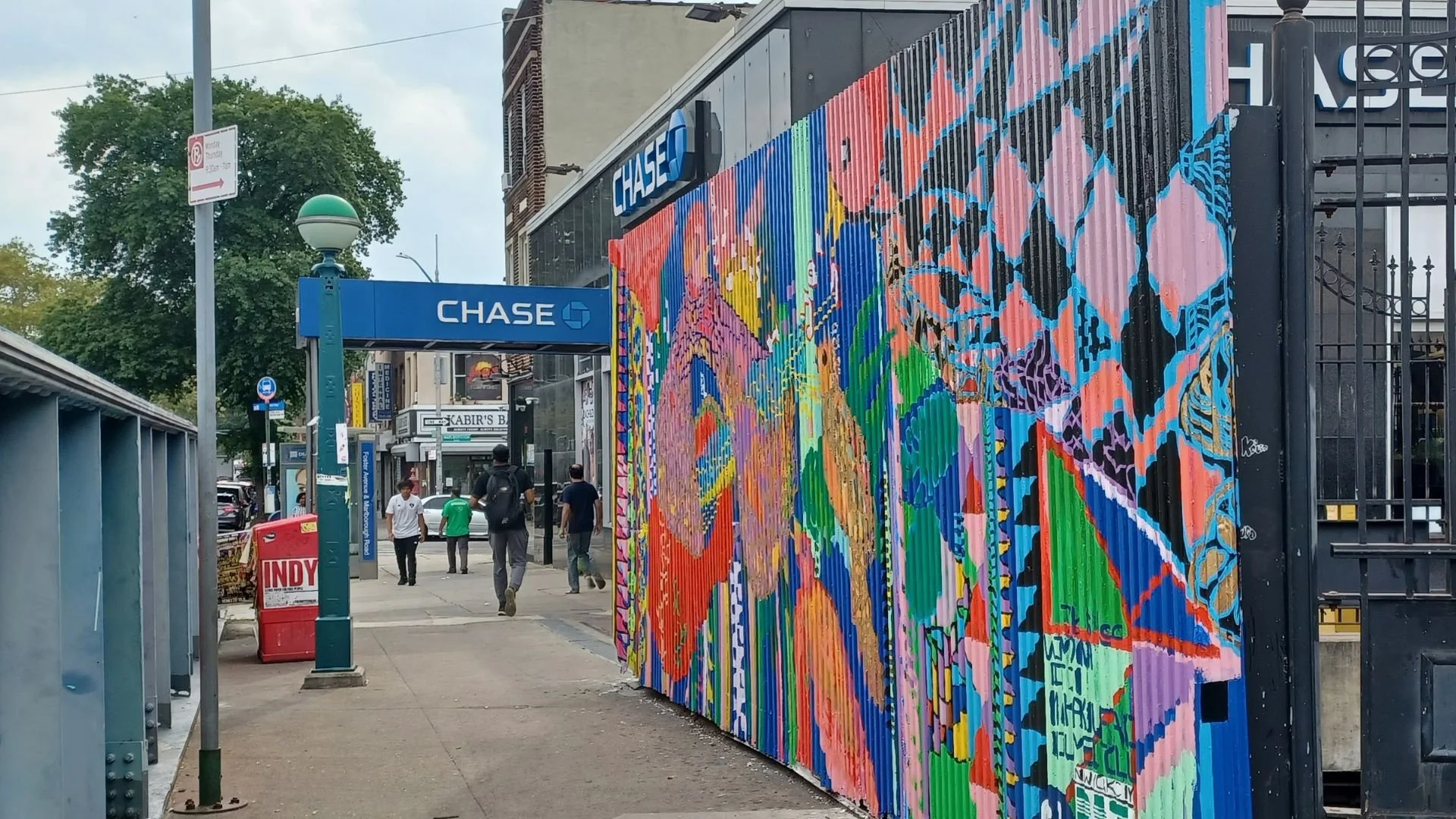 Street scene with a colorful mural on a black container, Chase bank sign, pedestrians walking, and storefronts in the background.
