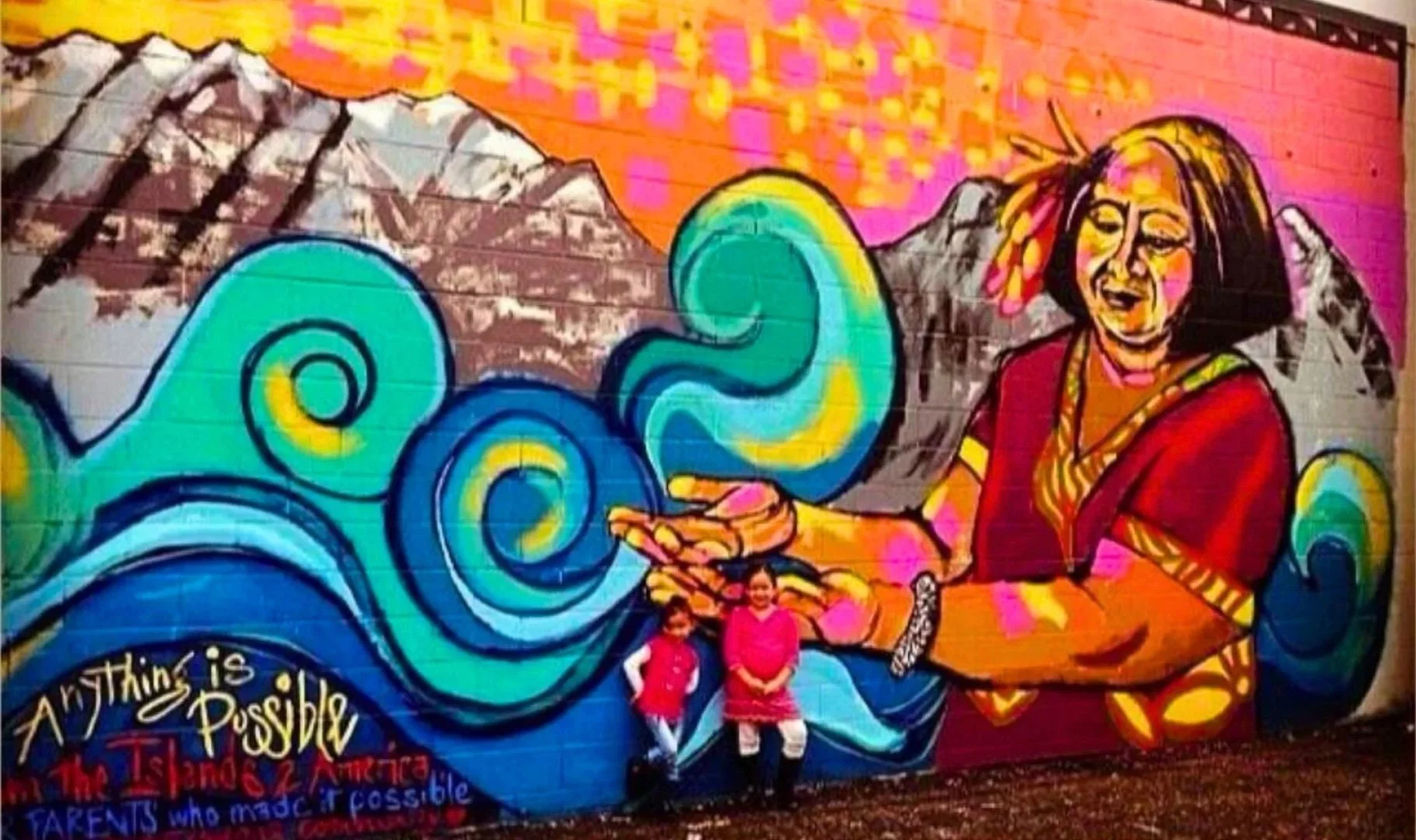 Colorful mural depicting a woman with short hair in traditional attire, extending her arm towards swirling blue and green patterns. Mountains are in the background, with pink and orange sky. Two children sit in front of the mural. The words "Anything is Possible" and a quote about parents and the American dream are painted at the bottom.