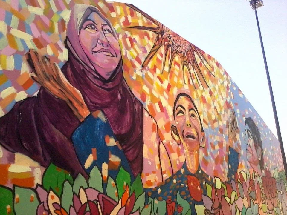 Colorful mural featuring diverse people, including a woman in a hijab, a crying young man, and other individuals, surrounded by vibrant patterns and floral designs.