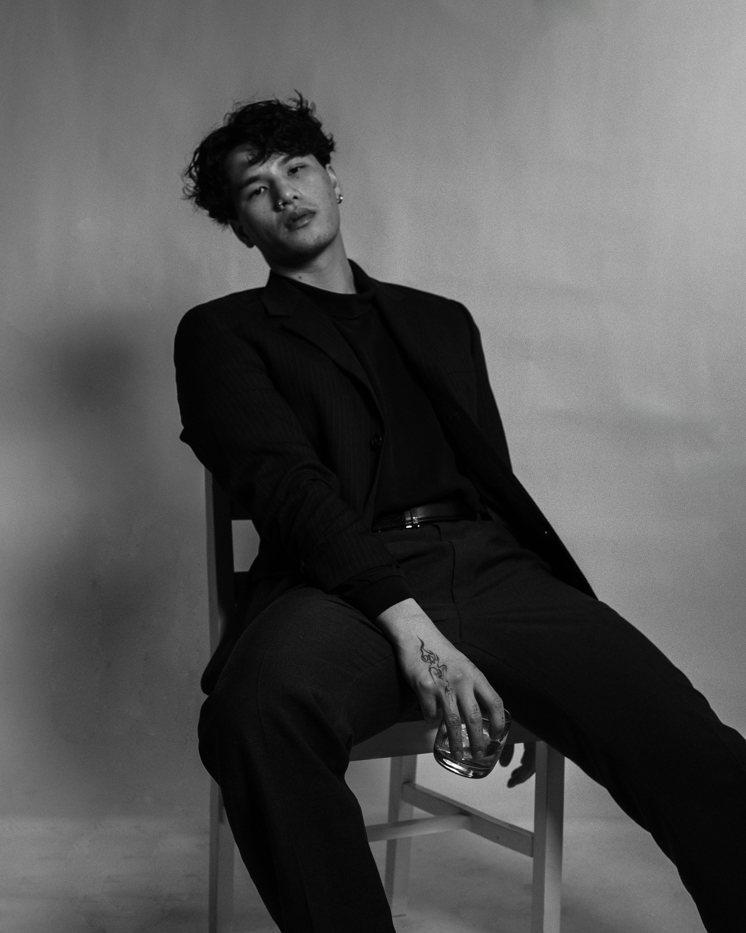 Black and white photo of a young man sitting on a chair, wearing a dark blazer and dark pants, holding a glass, with a relaxed pose and a neutral expression.