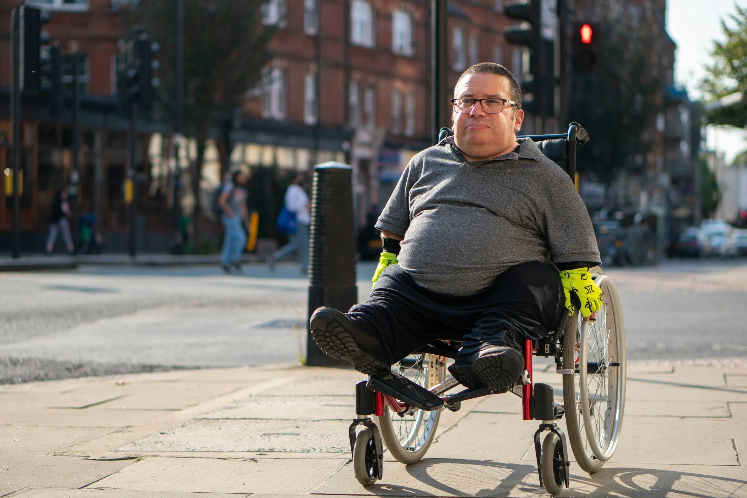 A man in a wheelchair sits on a city street corner, wearing glasses and a gray shirt, with buildings and pedestrians in the background.