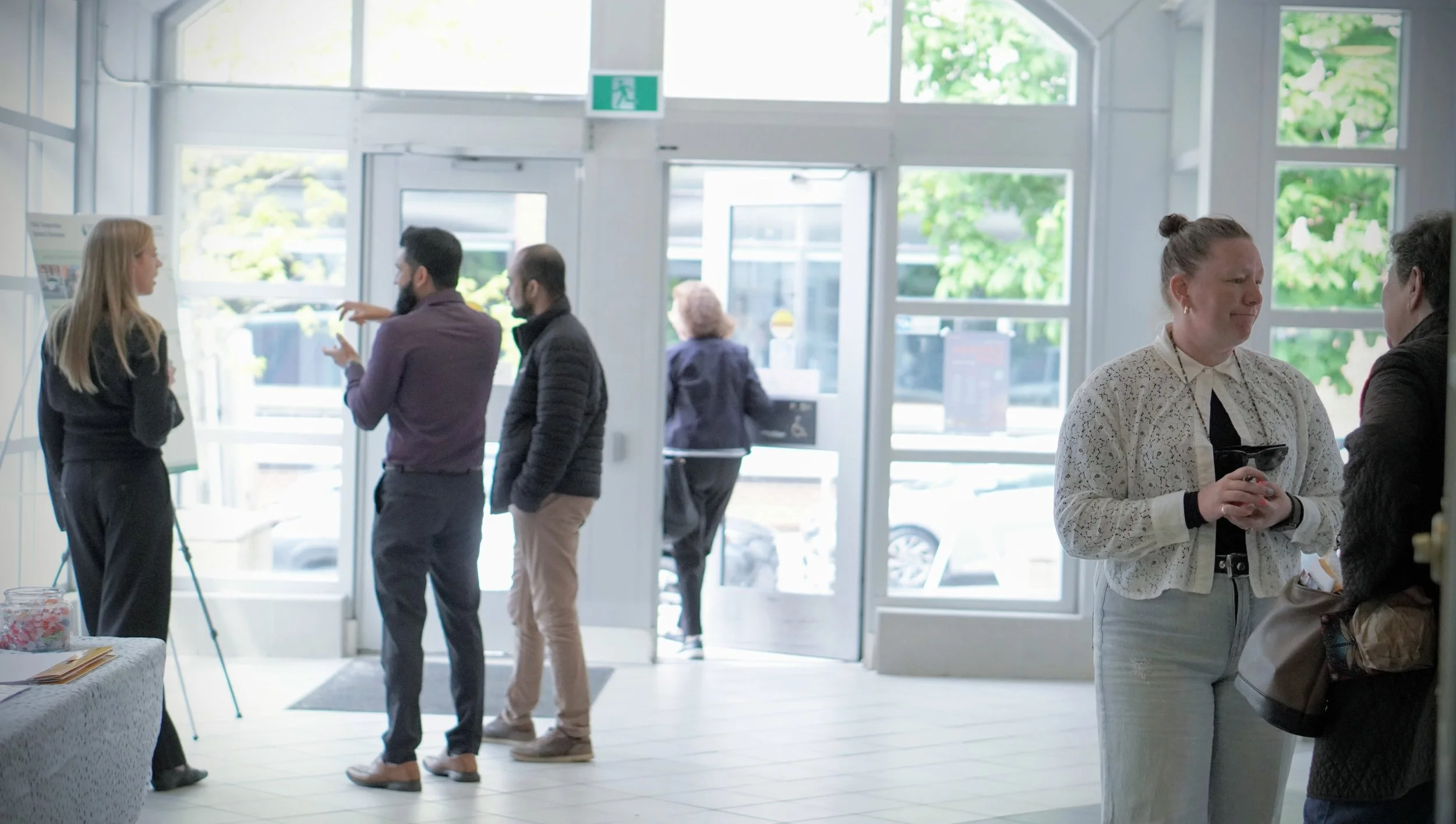 People standing and talking inside a bright building with large windows during a public engagement event, some near an entry door, with greenery visible outside.