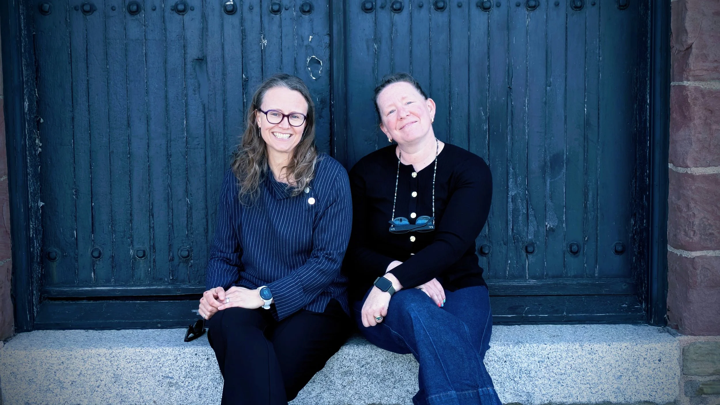 Kate and Claire are sitting on a bench in front of a dark wooden door. Kate has glasses, long wavy hair; Claire has her hair tied back, earrings. Both are dressed in dark clothing and are smiling at the camera.