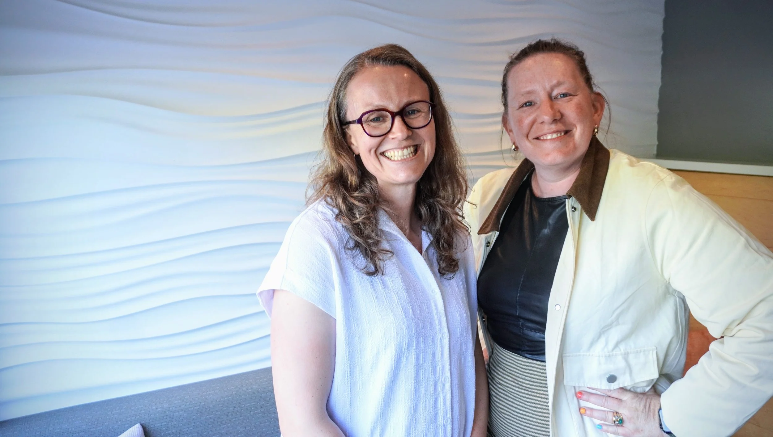 Claire and Kate are smiling for a photo inside a building with a textured wave-patterned wall in the background