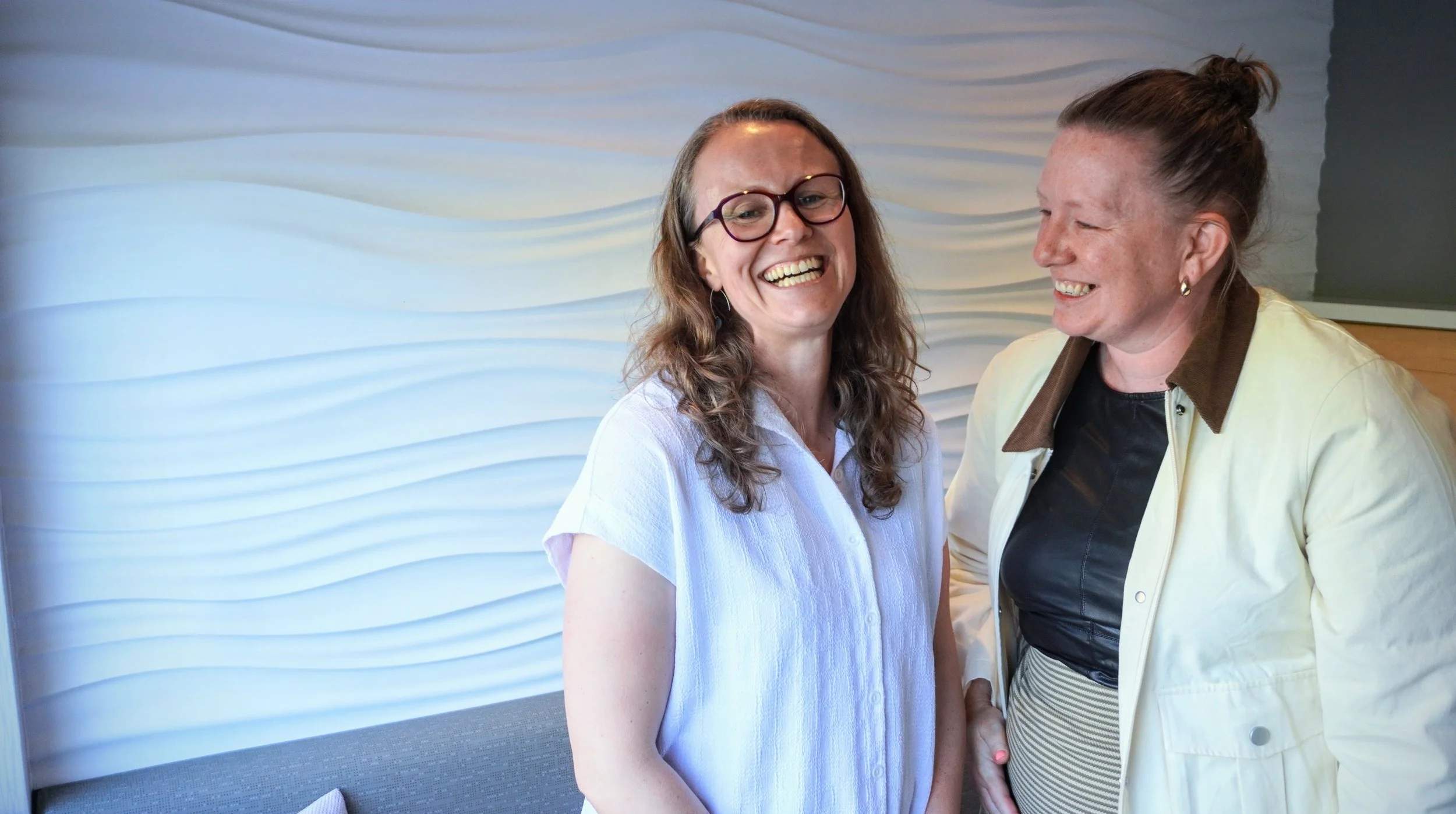 Kate and Claire are standing together, smiling and engaging in conversation indoors, with a textured, wavy white wall in the background.