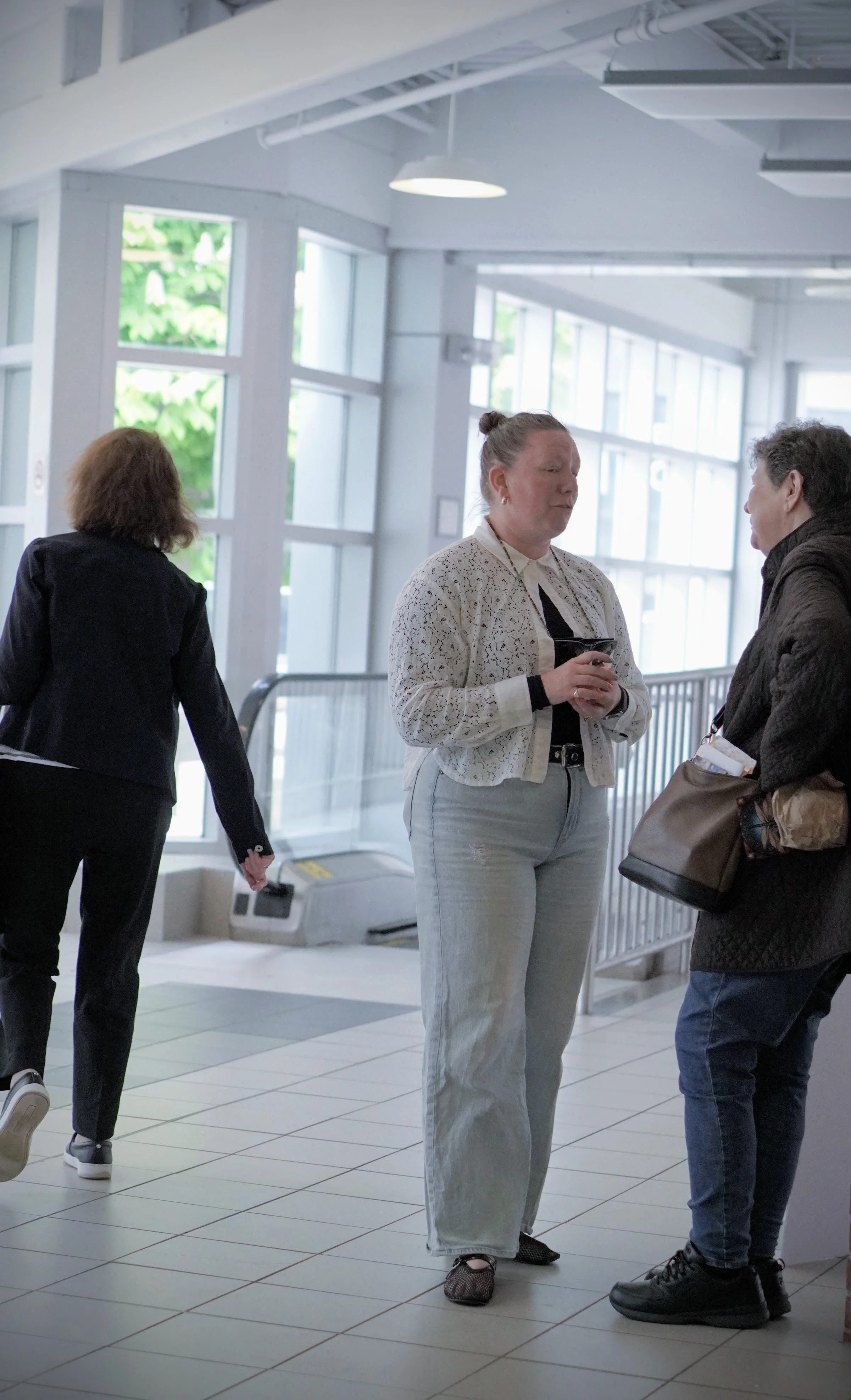 Three women standing and talking inside a building with large windows. One woman is facing the camera, holding a cup, wearing a white blouse and light gray pants. Another woman is facing her with short dark hair, wearing a dark jacket and holding a bag. The third woman is walking away, dressed in black.