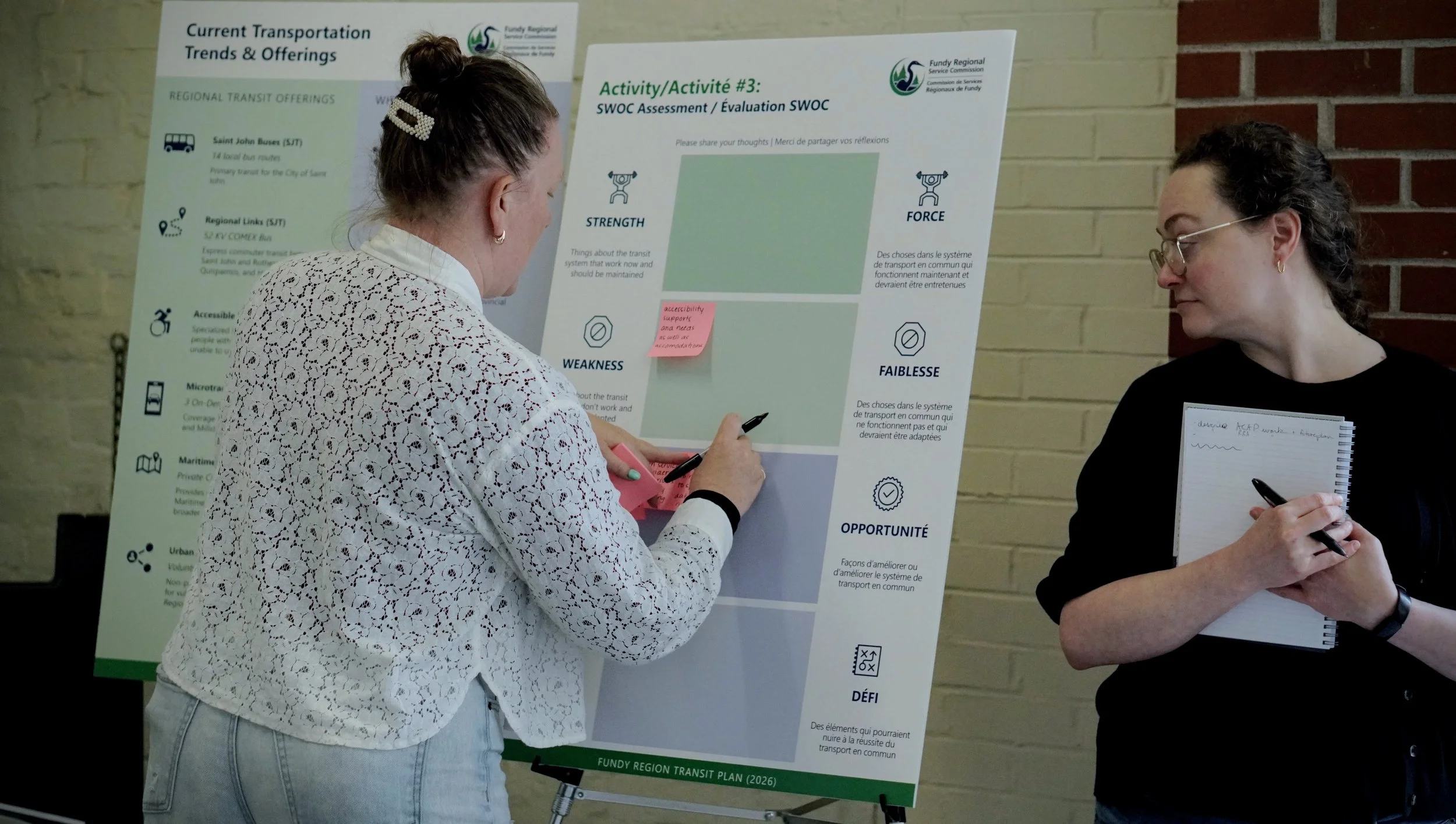Two people are standing by a board at a community engagement event. One person is adding sticky notes to the board.