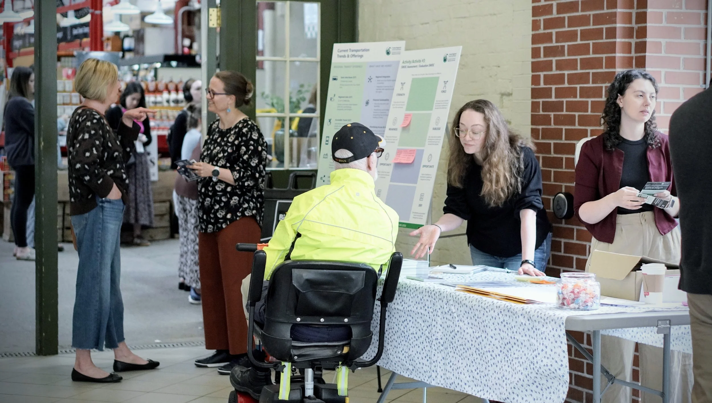 Two people are standing behind a table and talking to a person using a wheelchair whose in front of the table. Other people are standing and talking in the background.