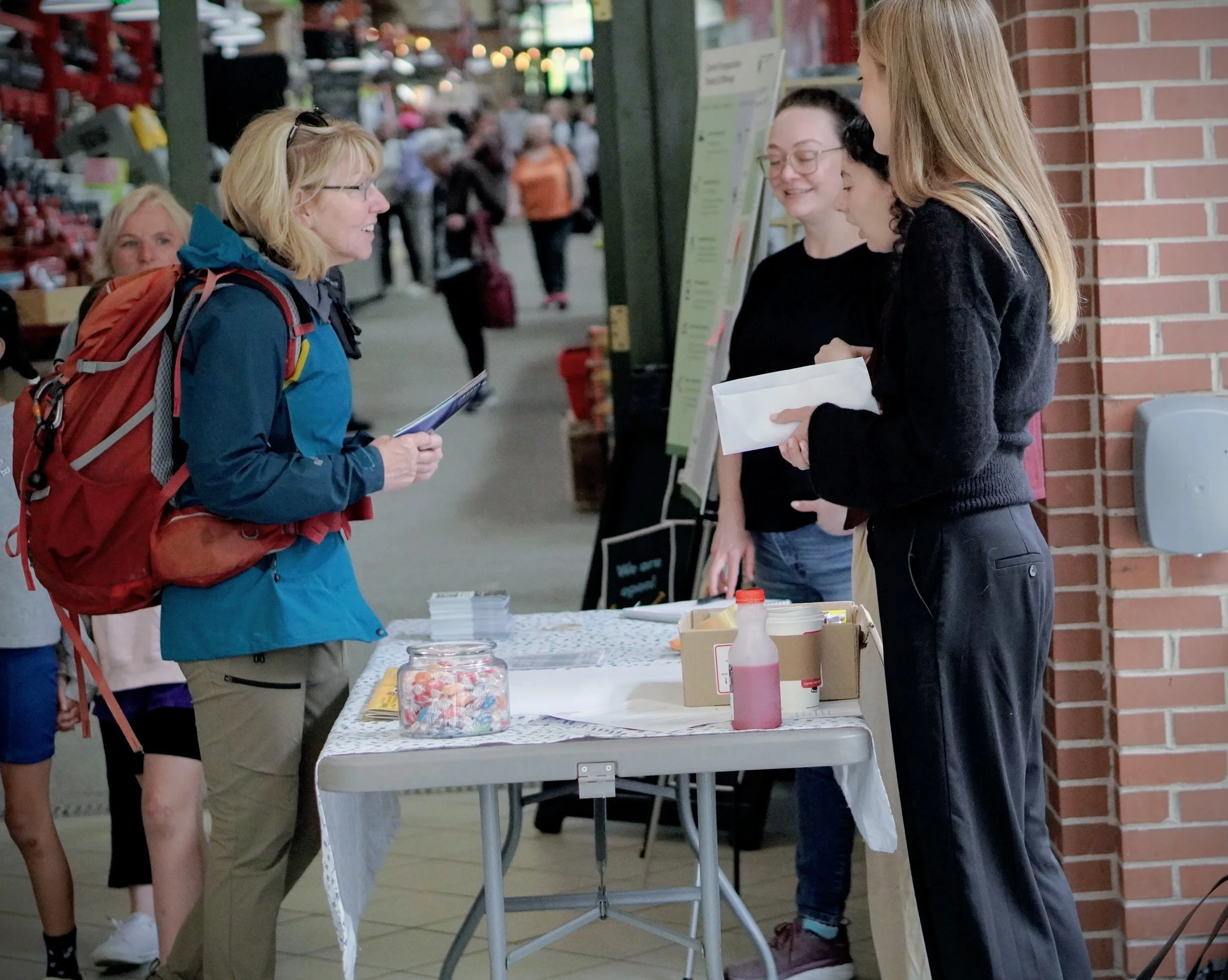A group of four people at an indoor public engagement event. They are interacting near a table with candy jars and pamphlets, with a crowd walking behind them.