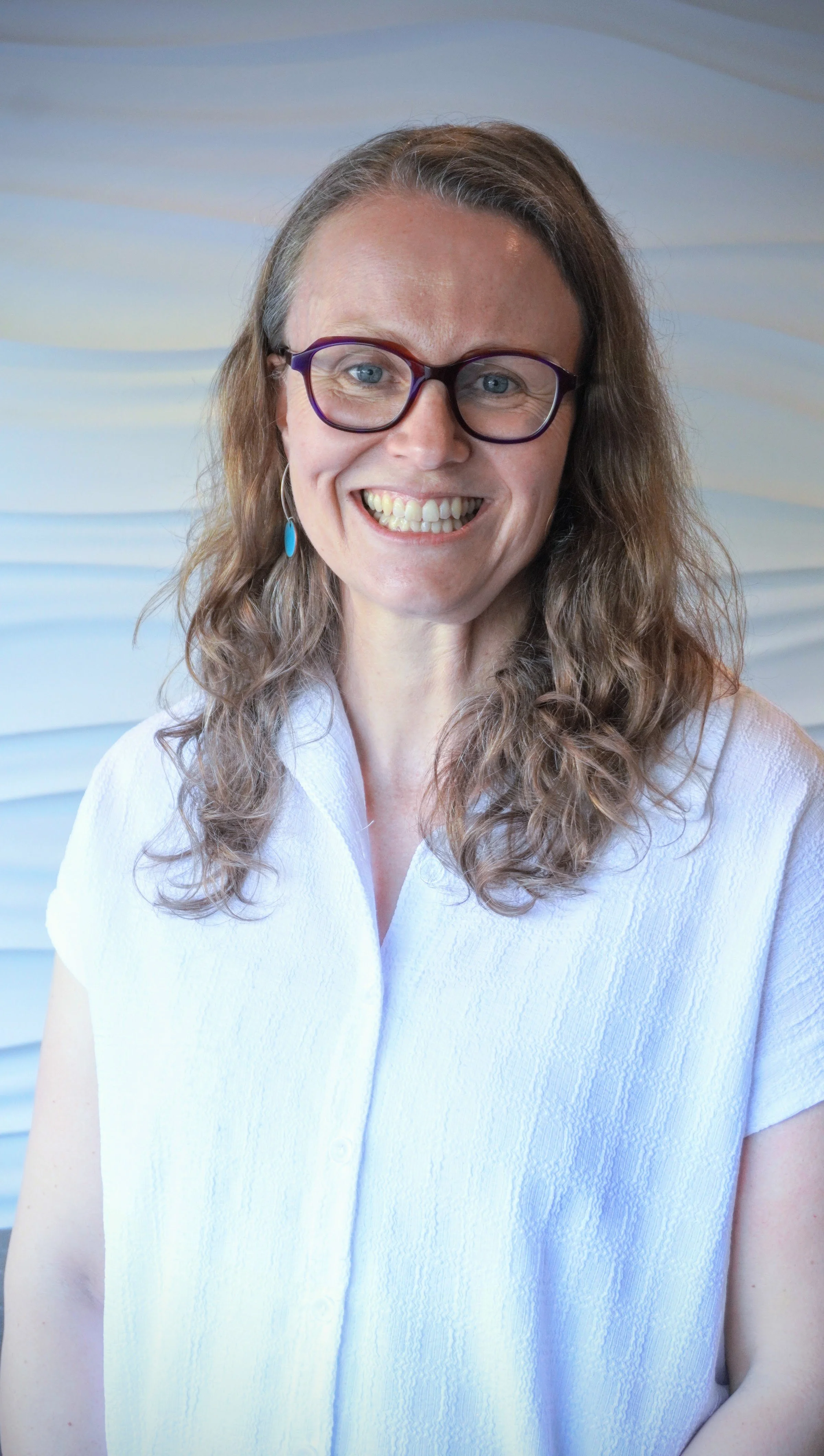 Kate, a woman with curly brown hair and glasses is smiling at the camera, wearing a white textured shirt, standing in front of a light blue background with horizontal wave patterns.