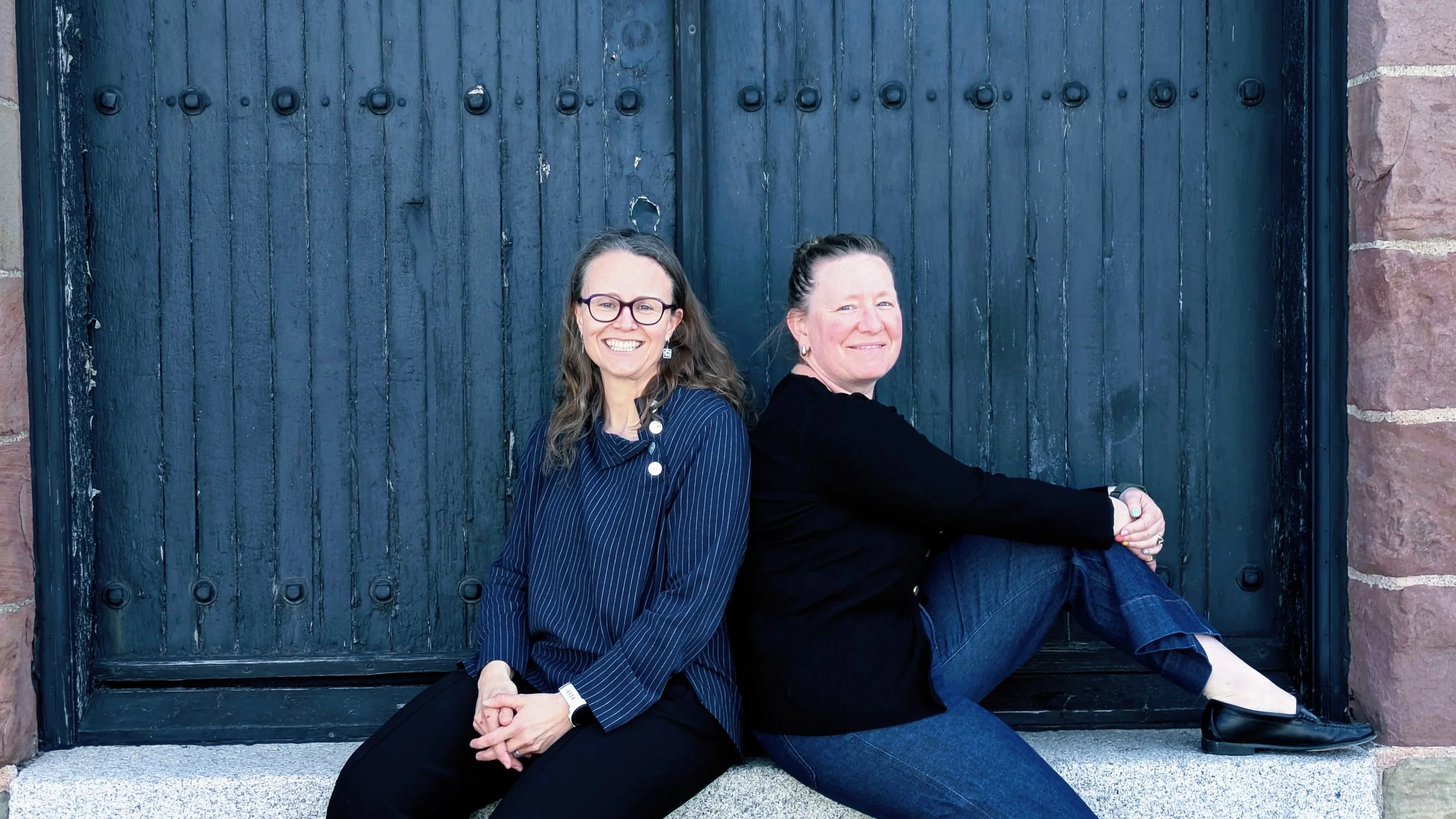 Kate and Claire, two consultants, are sitting together in front of an old wooden black door