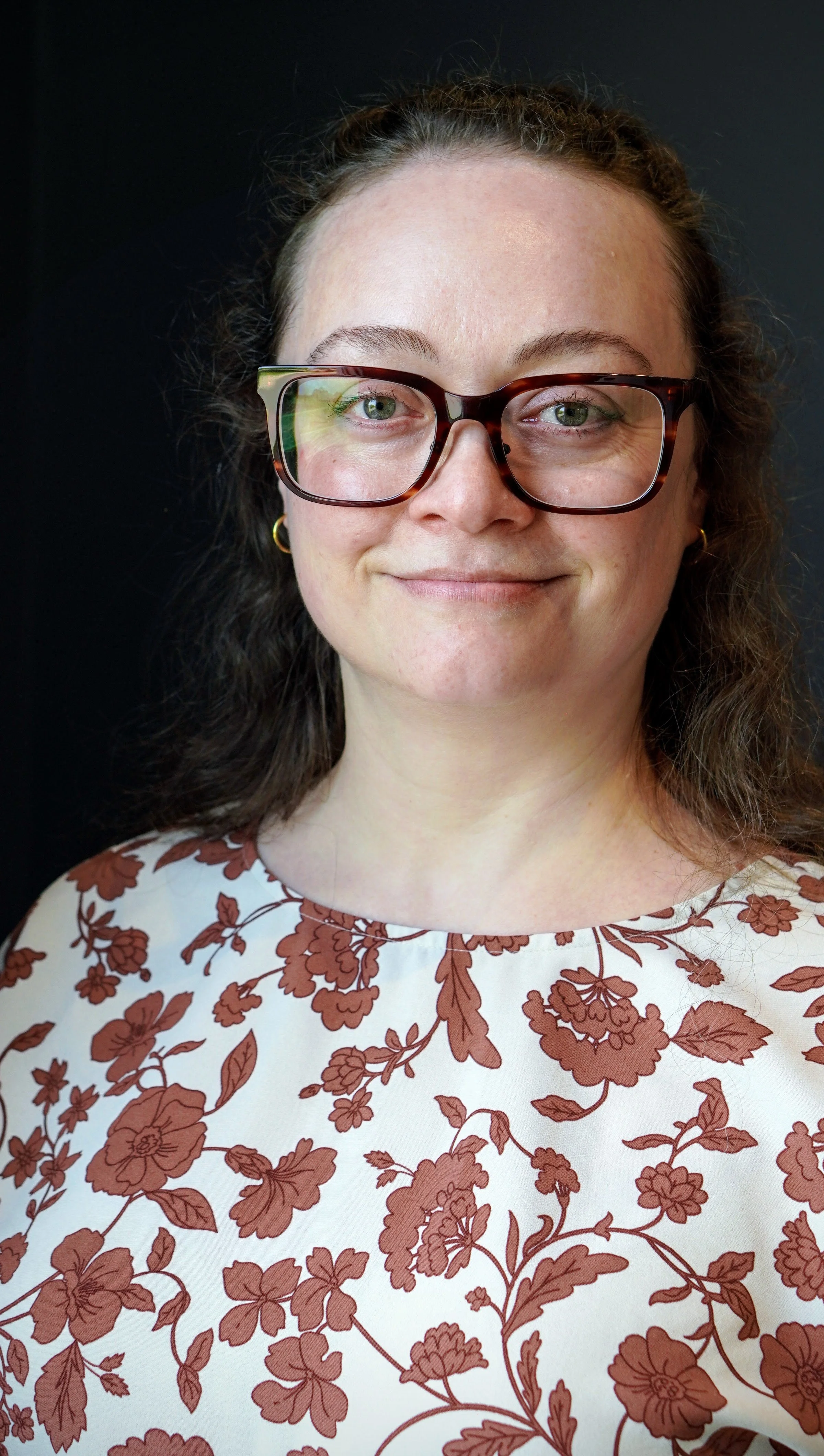 Samantha has curly brown hair and she's wearing large tortoiseshell glasses and a white dress with brown floral pattern. She's standing against a black background.