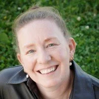 Claire has light hair tied back, wearing a dark jacket, and she's outdoors with greenery in the background.