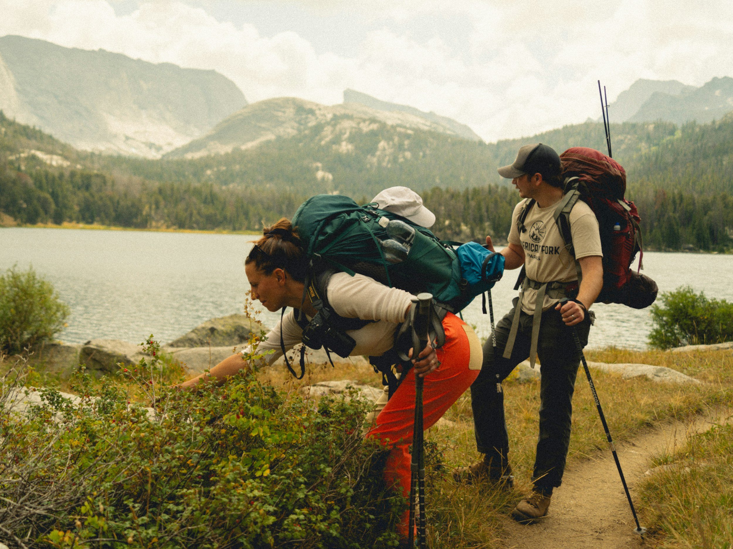 Two hikers in outdoor gear walking along a lakeside trail with mountains in the background.