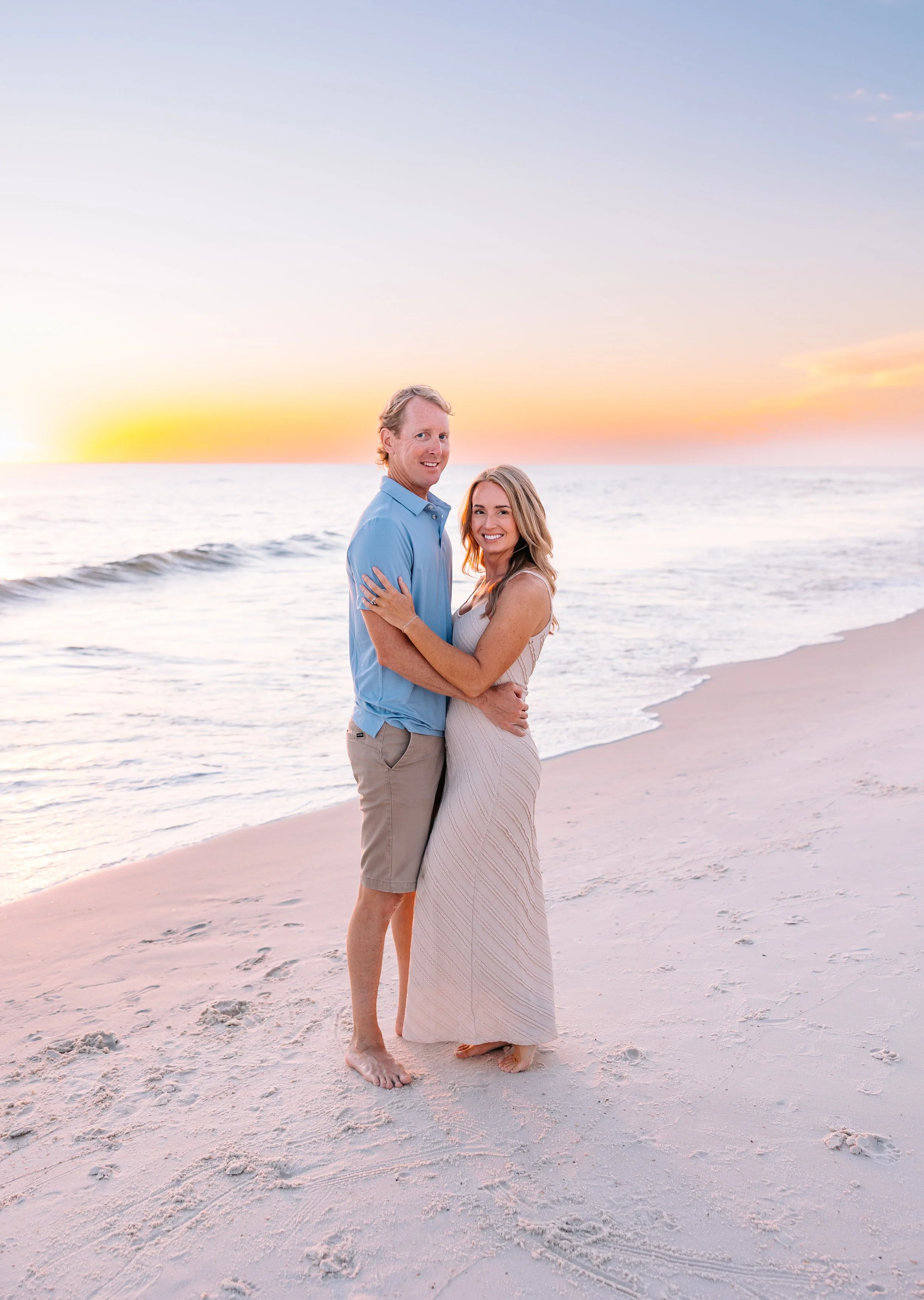 A couple standing on a sandy beach at sunset, with the man wearing a light blue shirt and beige shorts and the woman in a white dress, both smiling and embracing each other.