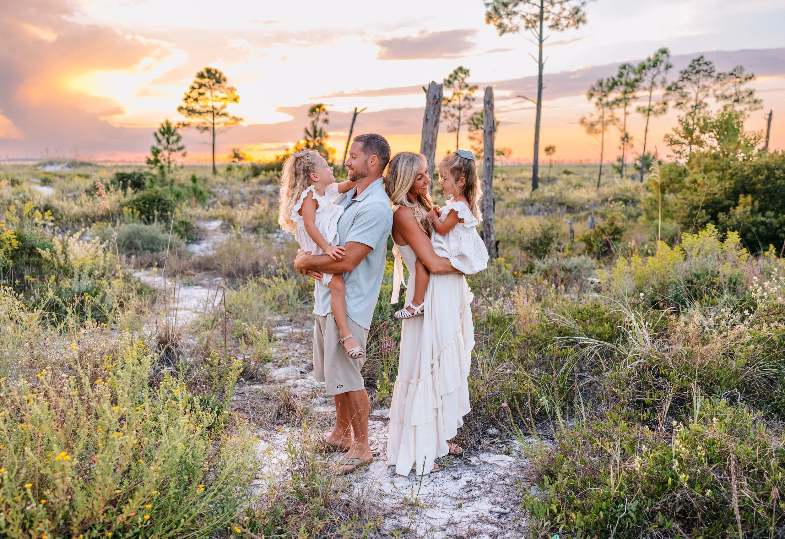 A family of four outdoors during sunset, with two adults and two young girls dressed in white, standing in a grassy area with trees and a partly cloudy sky in the background.