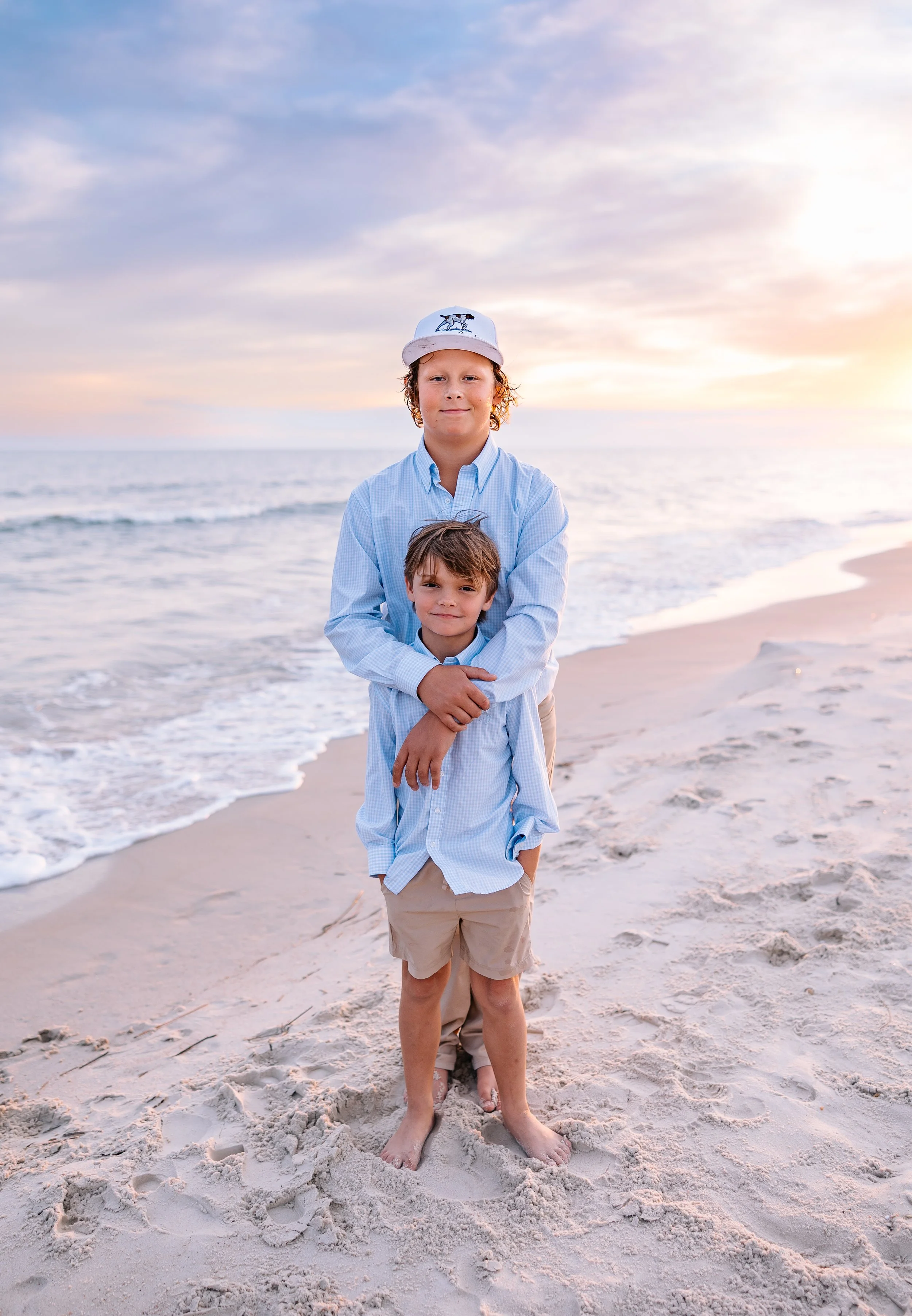 Two young boys with light skin and brown hair standing on a sandy beach during sunset, with the ocean and a cloudy sky in the background. The older boy, wearing a white cap and light blue shirt, stands behind the younger boy who is in a light blue shirt and khaki shorts. They are barefoot in the sand.