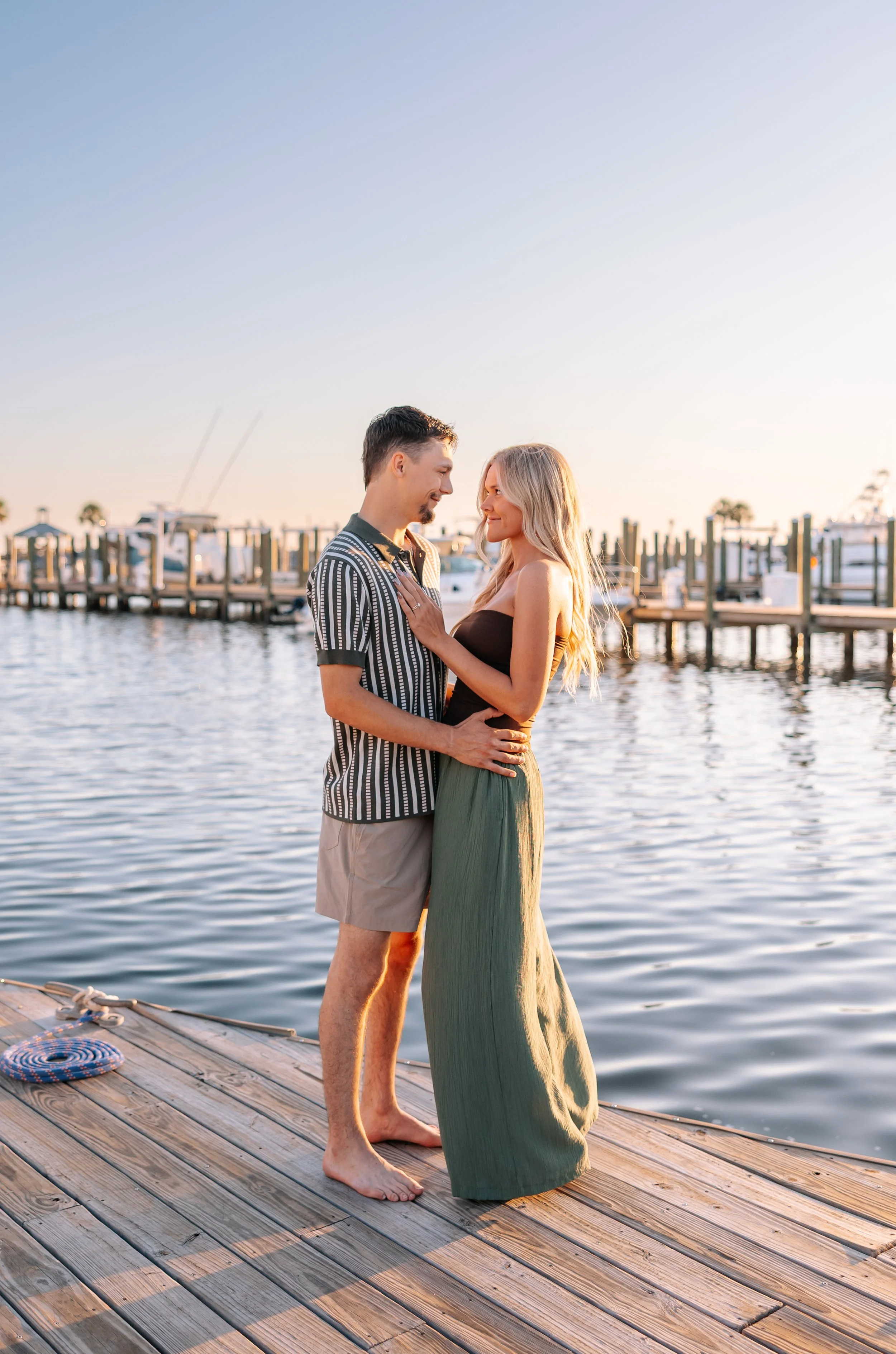 A couple stands close together on a wooden dock by the water during sunset, facing each other with smiles, with boats and a marina in the background.