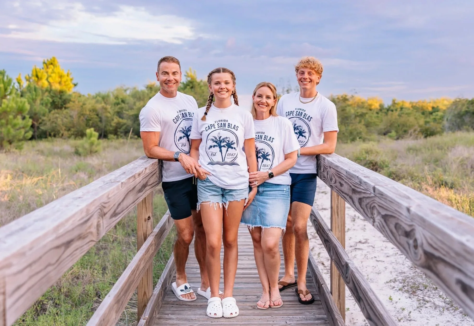 A group of five people, two men and three women, standing on a wooden bridge in a natural outdoor setting with trees and grass in the background. They are smiling and wearing matching white t-shirts with a palm tree graphic and the text 'Florida Cape San Blas Est. 1847'.