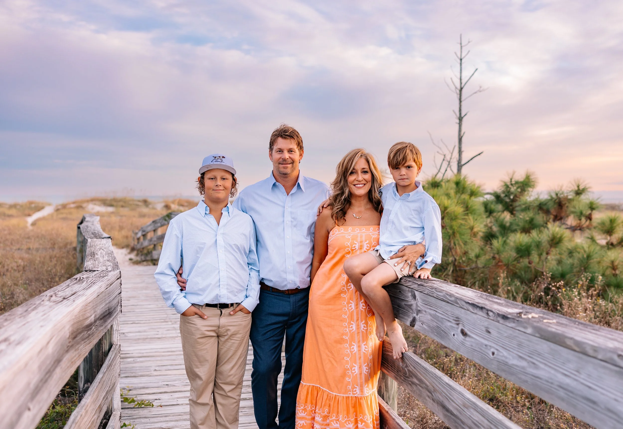 A family of four standing on a wooden boardwalk in a natural, outdoor setting during dusk. The family includes a man, a woman, and two boys. The background features a cloudy sky, a dry, grassy landscape, and sparse vegetation.