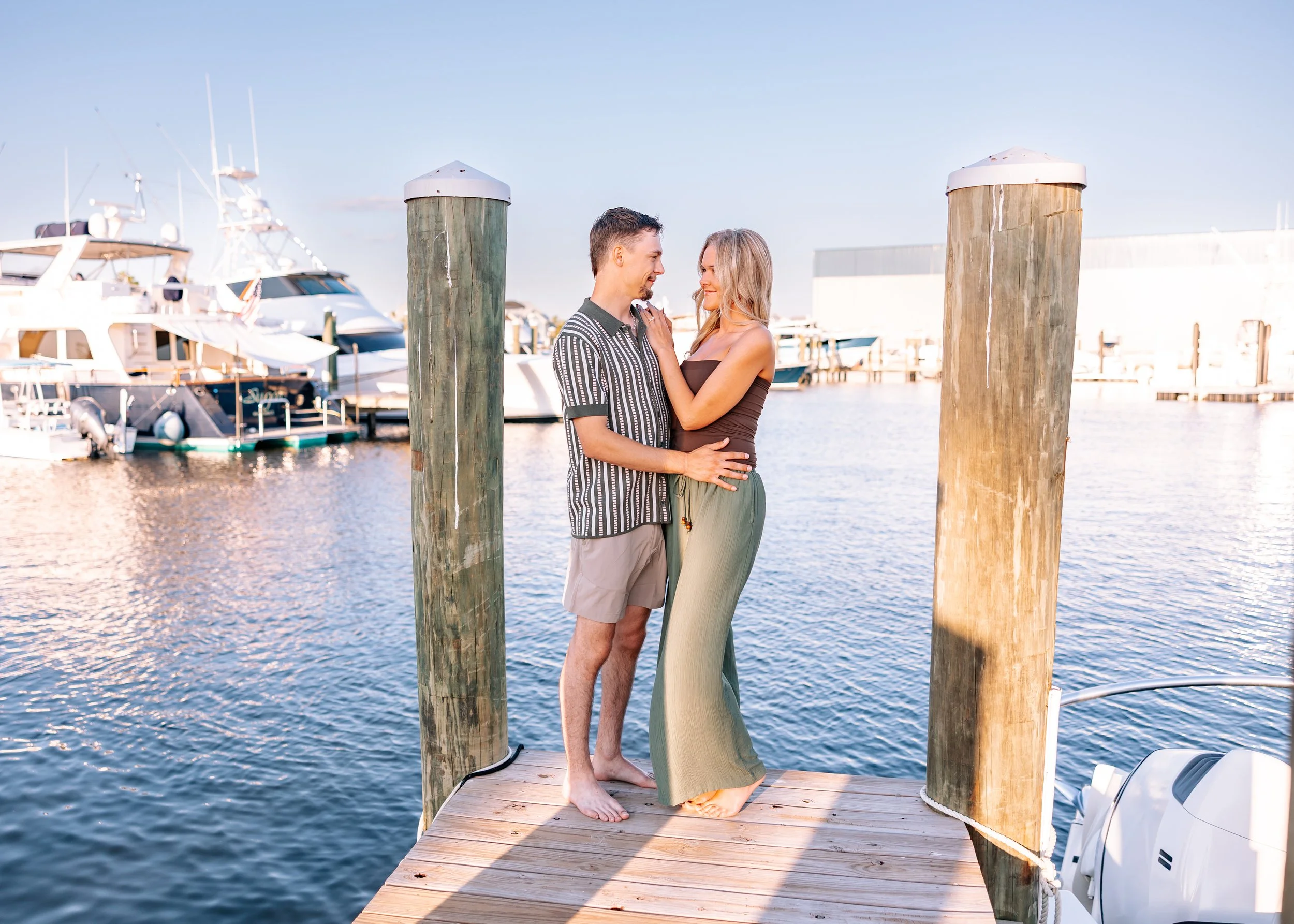 A couple standing on a dock by the water, with yachts in the background, smiling and looking into each other's eyes.