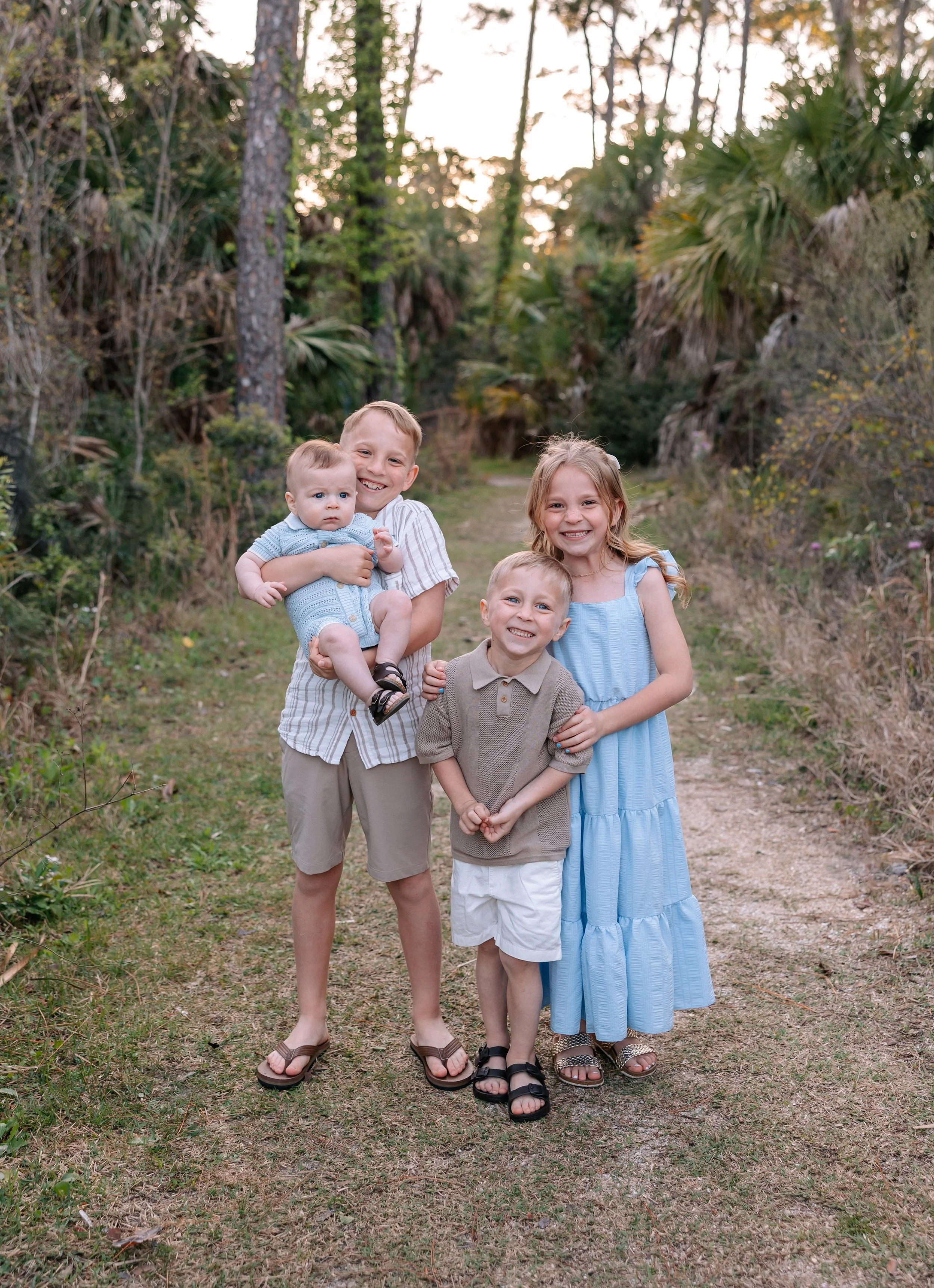 Group of four children, two boys and two girls, standing on a dirt path in a wooded area, smiling, with trees and bushes in the background at sunset.