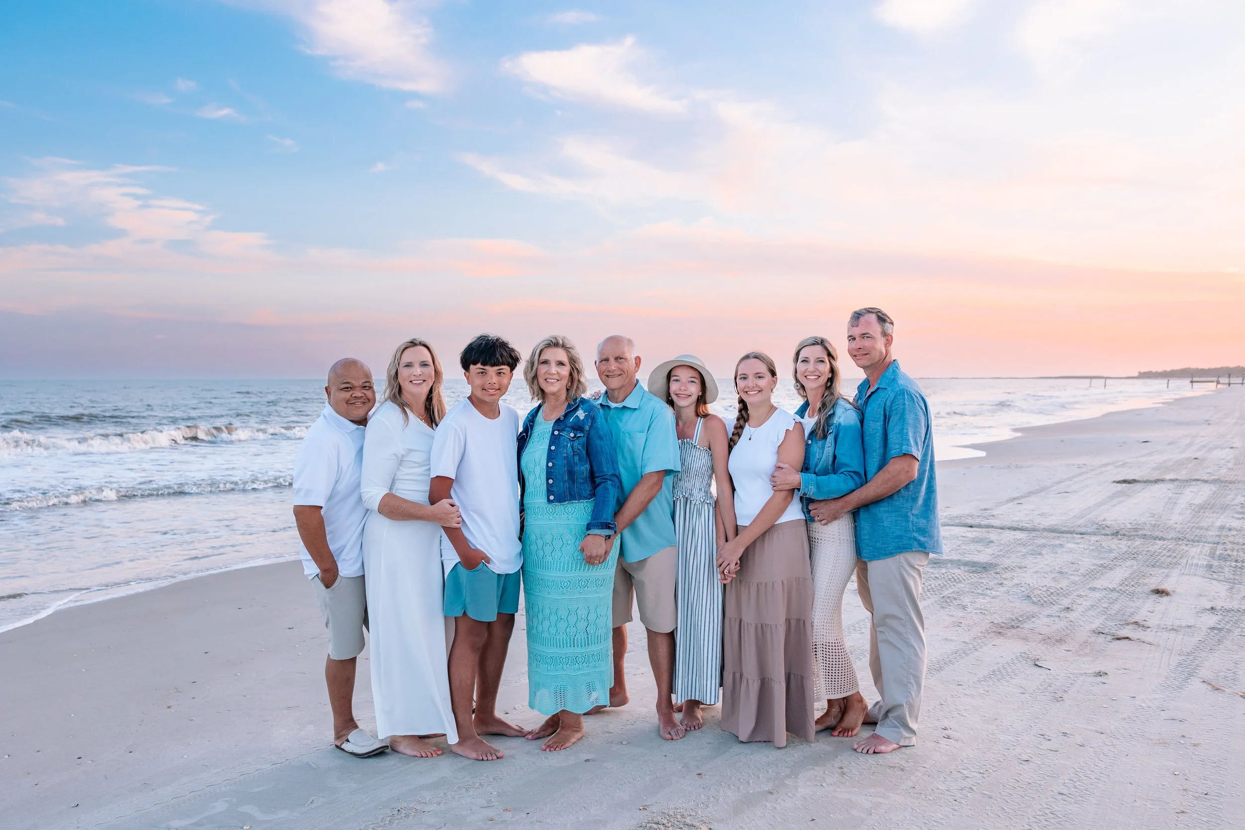 A diverse group of ten people, including children and adults, standing on the beach at sunset, smiling and dressed in casual, summery clothing.