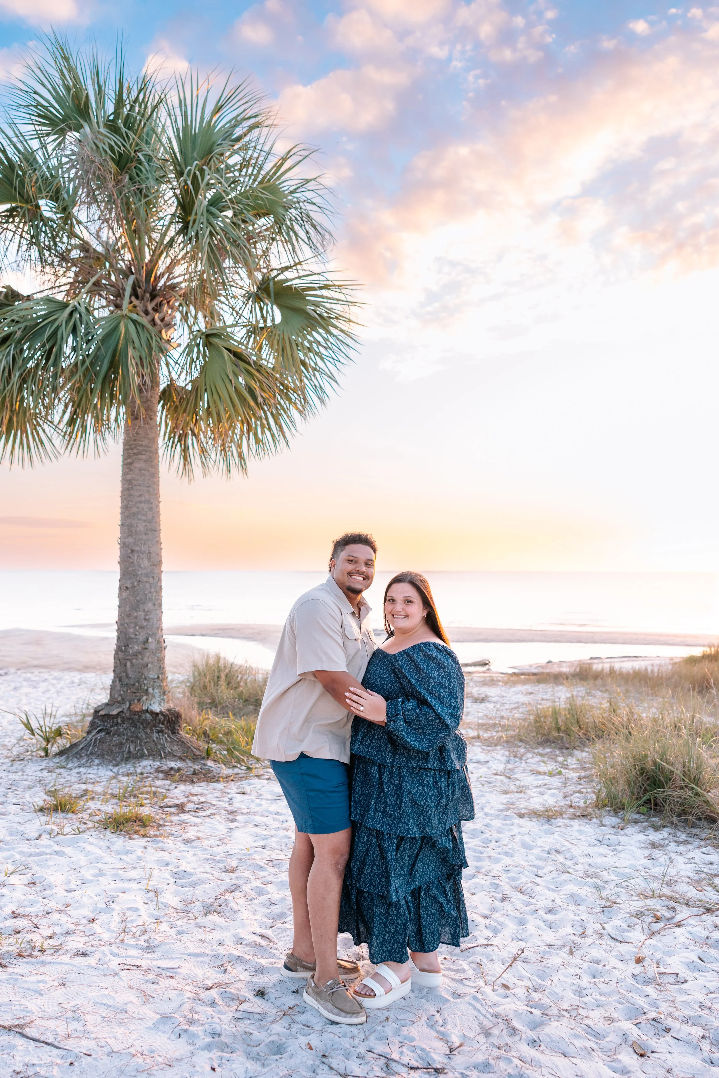 A smiling couple standing on the beach at sunset, with a palm tree on the left and a colorful sky with clouds.
